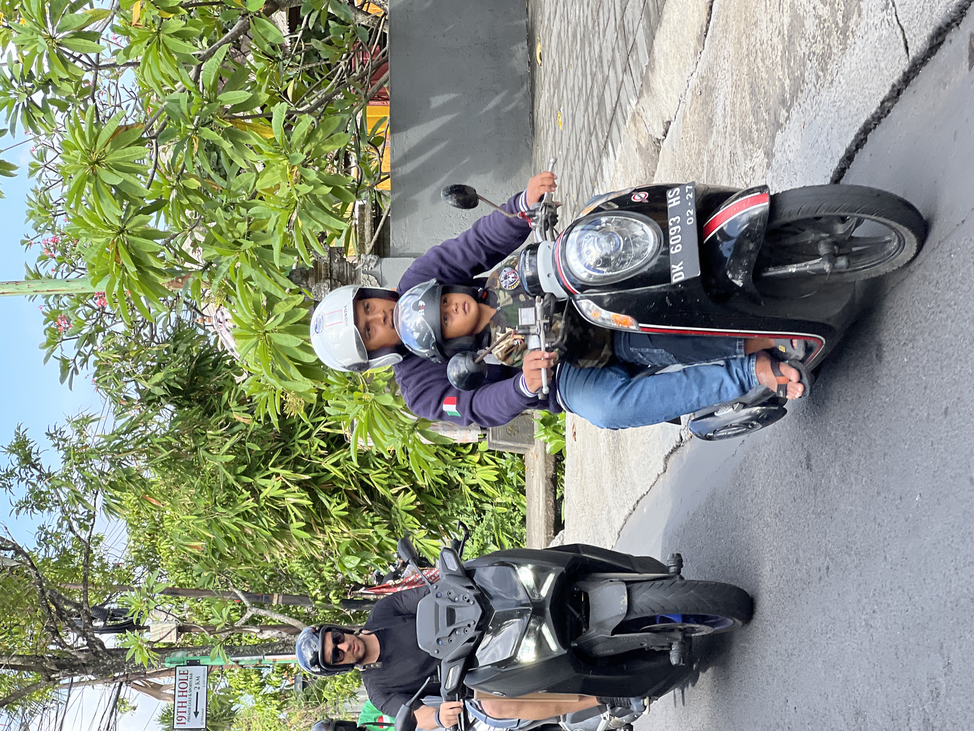 In North Kuta, Indonesia, on a paved road, two individuals are riding on separate motorcycles. The foreground features a black scooter with a rider wearing a white helmet and a dark blue jacket with an Italian flag patch on the left sleeve. A child, wearing a dark helmet and a camouflage jacket, is seated in front of the rider. The scooter's license plate reads "DK 6093 HS" and "02-27". To the left, a black motorcycle carries a rider with a black helmet and sunglasses, wearing a black t-shirt and blue shorts. The background displays lush green trees, foliage, and a sign that reads "19TH HOLE PREMIER GOLF & SPORTS BAR 2 KM". The sky is clear and blue.
