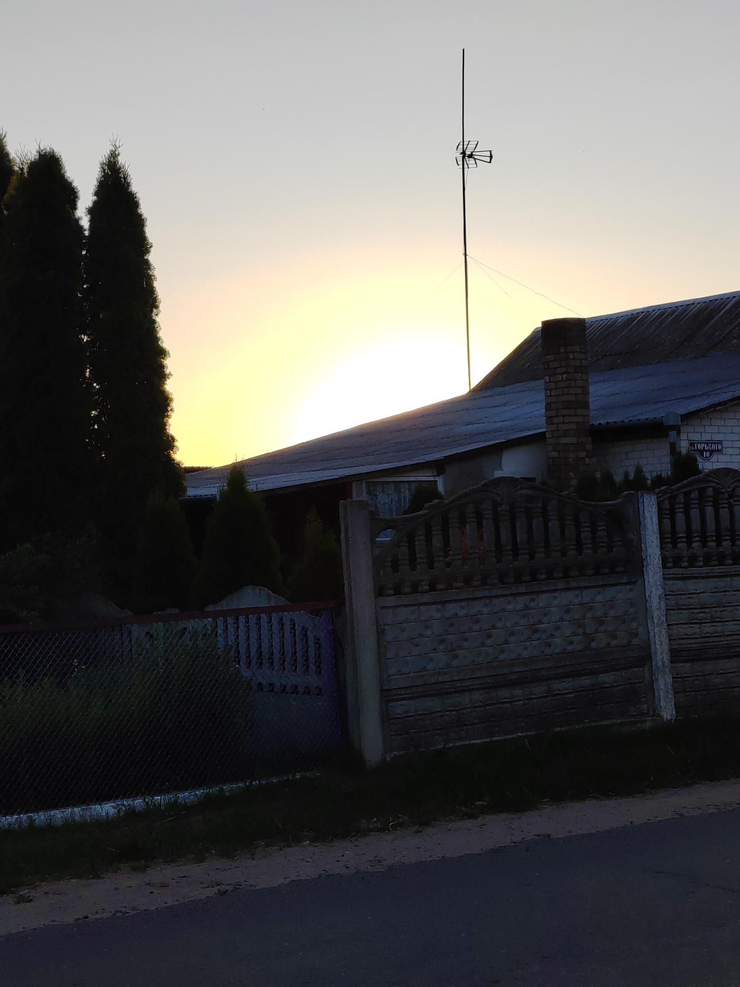 This image depicts a rural scene at dusk in the Воўчынскі сельскі Савет, Belarus. The setting sun casts a warm, yellow glow across the horizon, partially obscured by the dark silhouettes of several tall evergreen trees on the left side of the frame. Behind the trees and the fence, a building with a dark, sloped roof and a brick chimney is visible. An antenna on a tall pole stands against the sky above the building.
In the foreground, there is a chain-link fence and a section of a decorative concrete fence with ornate posts and horizontal planks. A dark asphalt road runs in front of the fence, with a strip of grass and dirt between the road and the fence.
A street sign with Cyrillic text is mounted on the side of the building. It reads "ГОРЬКОГО 10". The overall mood is peaceful and serene, capturing the quiet end of a day in the countryside. The time of day is clearly indicated by the low position of the sun and the soft, diffused light. There are no people or animals visible in the frame. The weather appears clear, with a pale sky.