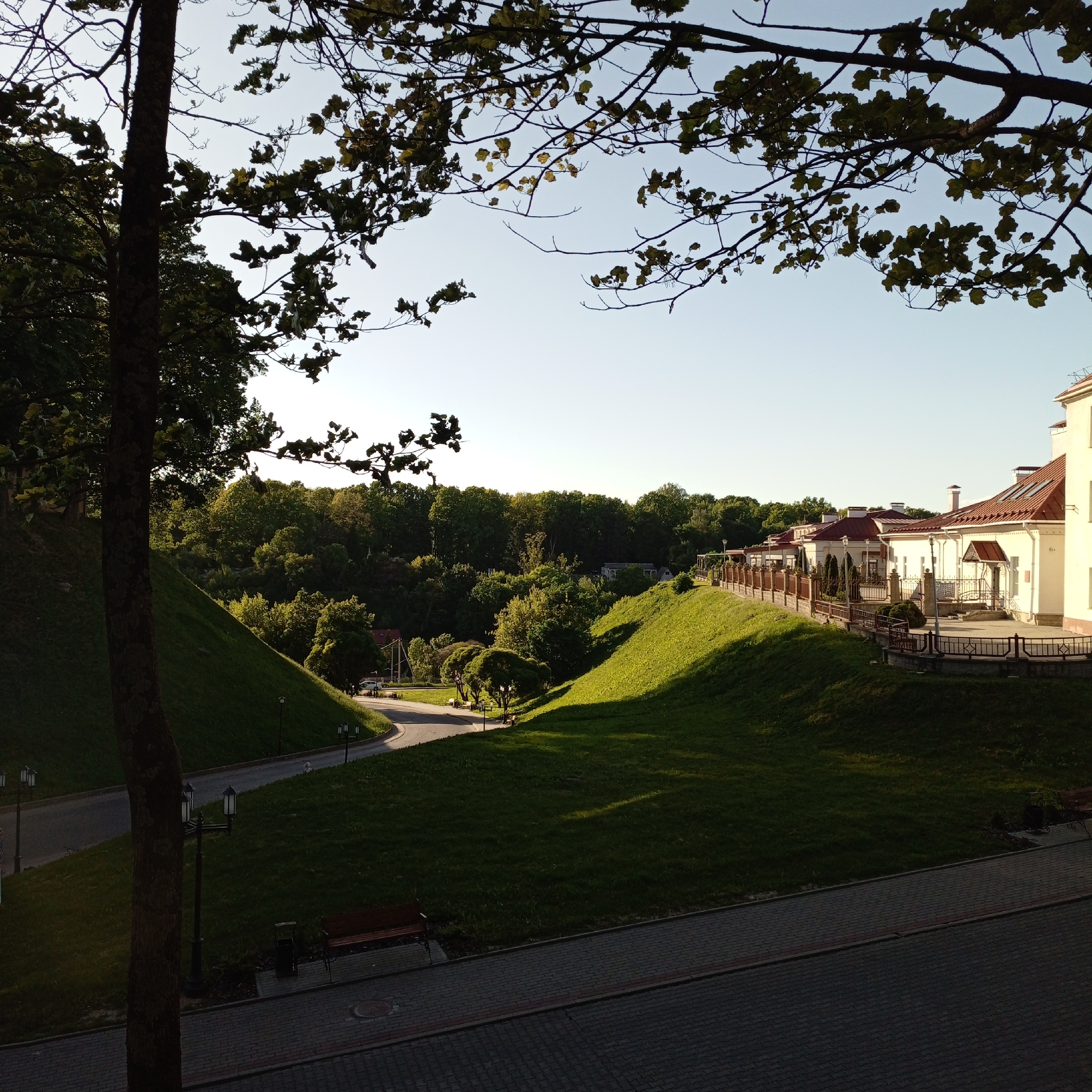 The image shows a landscape with a road curving between grassy hills. On the right side, a building complex with red tile roofs stands atop one of the hills, enclosed by a fence and greenery. The lighting suggests late afternoon or early morning, with shadows cast across the grass. In the foreground, there is a paved area with a bench and streetlights. Trees frame the scene at the top and left. The location is Unknown, Unknown.