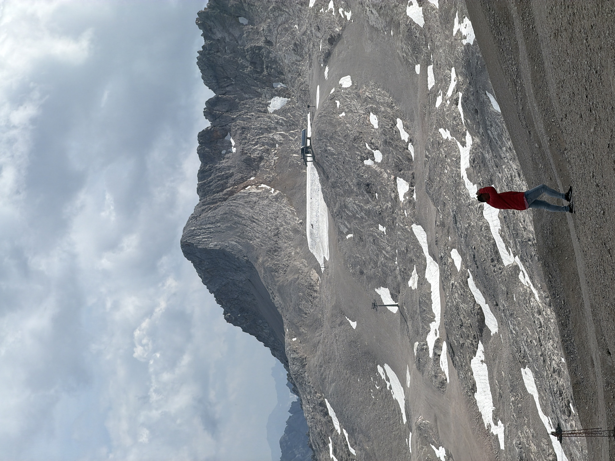 A person in a red jacket and blue jeans walks across a barren, rocky slope dotted with patches of snow. The person is facing away from the camera, with their hands raised to their head. In the background, a large, rugged mountain range dominates the scene, with a ski lift and a small building visible on a plateau near the summit. The sky is overcast with dramatic clouds. This image was taken at the Zugspitze, Germany's highest peak, near Garmisch-Partenkirchen.