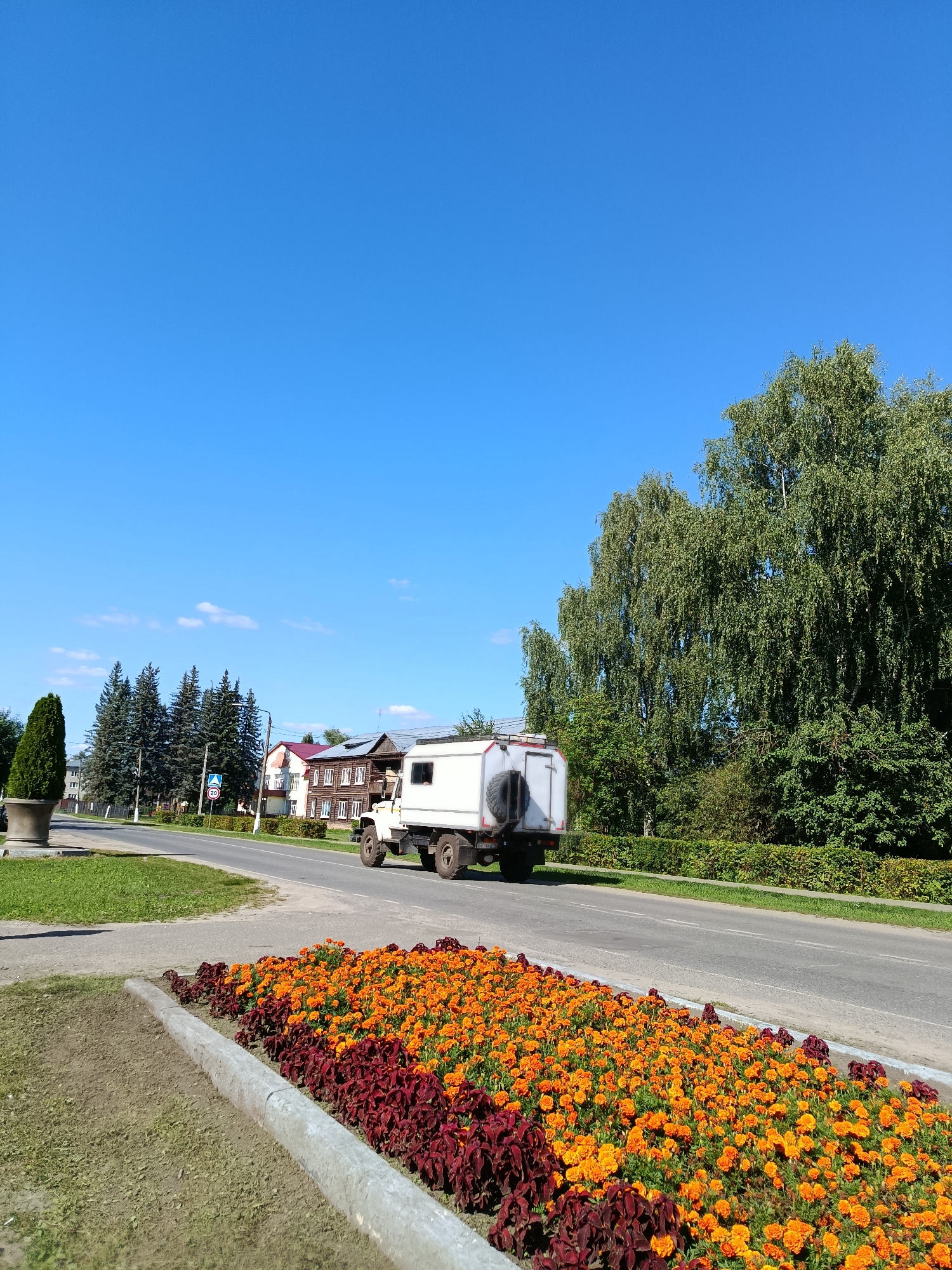 A white utility vehicle with a camper shell is parked on a paved road in a town. The vehicle has a spare tire mounted on the rear. A line of buildings, some appearing to be wooden, are visible across the road. Large trees, including birches, line the right side of the road. In the foreground, a flower bed with orange and maroon flowers is visible. A traffic sign indicating a speed limit of 20 is posted on the left side of the road. The sky is clear blue with a few small white clouds. The scene is consistent with a daytime setting in the town of Vereya, Russia.