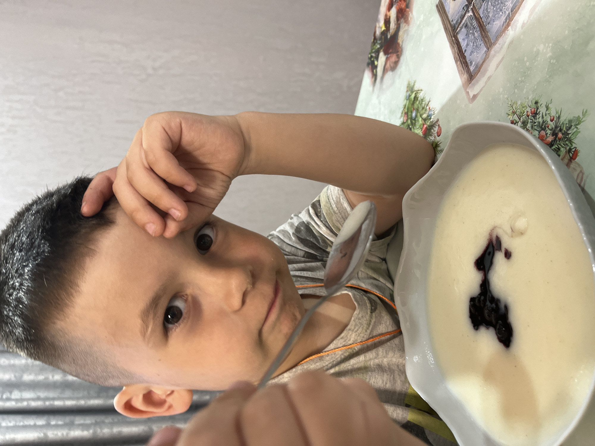 A young boy sits at a table in Chaikovsky, Russia. He holds a spoon in one hand and rests the other on his forehead. In front of him is a bowl of white porridge with dark jam in the center. The table is covered with a patterned tablecloth, and the background shows a white wall.