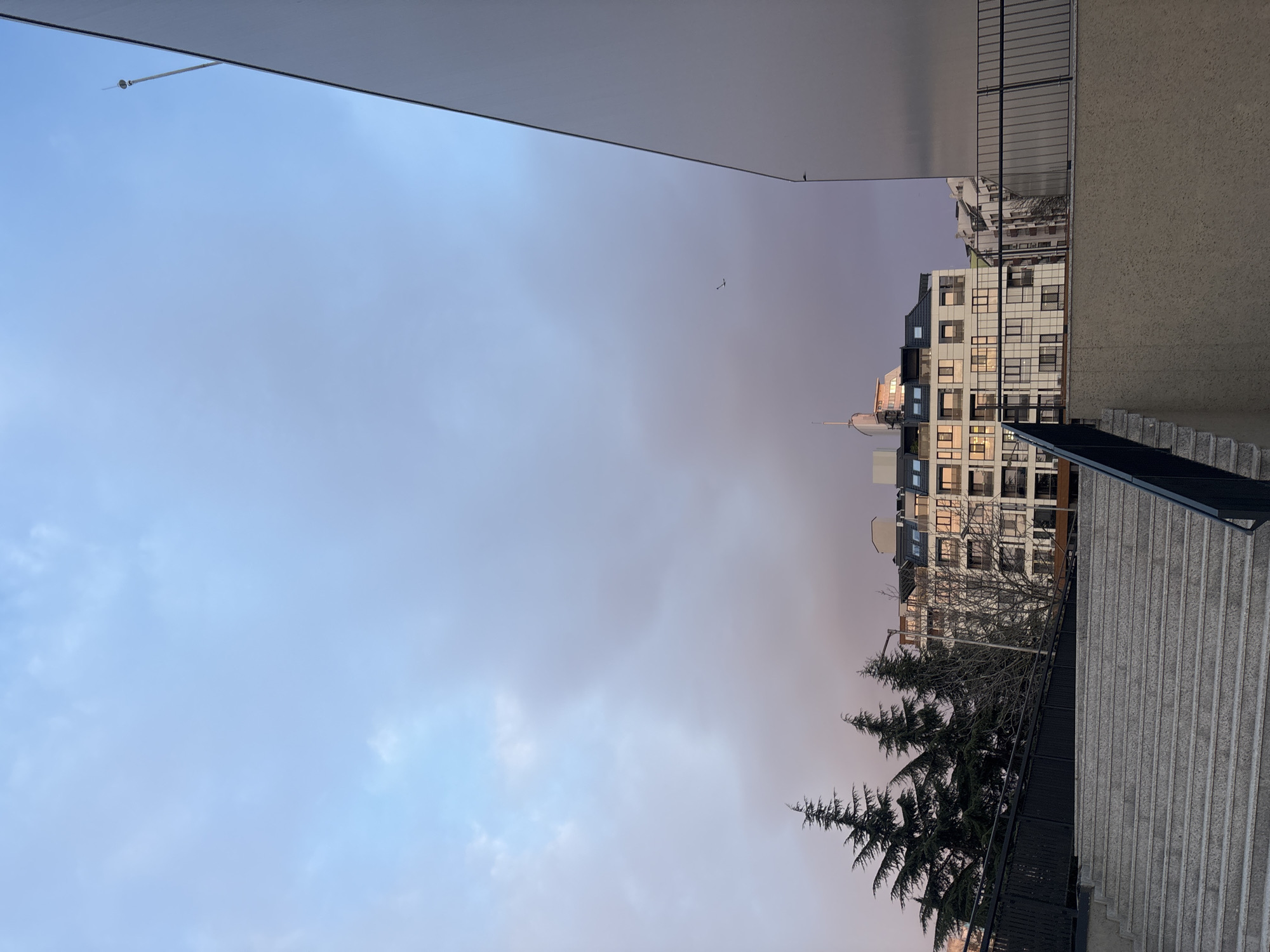 The photograph shows an urban scene, likely in Burgas, Bulgaria, given its architectural style. The image is taken from a slightly elevated position, possibly a building or a set of stairs. To the right, a concrete staircase with black metal railings descends. Adjacent to the stairs is a tall, multi-story building with a modern facade, featuring many windows. A few bare tree branches and evergreen trees are also visible near the building. In the sky, a light blue hue is present on the left, transitioning to a pinkish-purple towards the right, suggesting either dawn or dusk. A bird is flying in the sky. The top of the frame is dominated by a light-colored building structure that seems to be part of the same building.