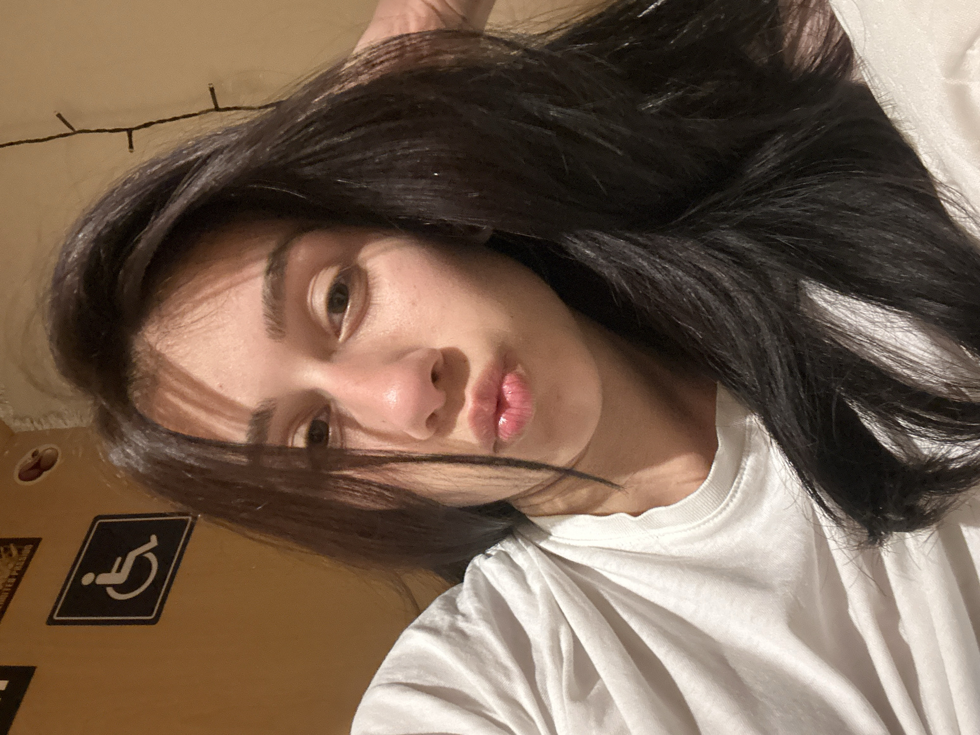 A low-angle, close-up selfie of a young woman with long, dark hair wearing a white t-shirt. The woman has light skin, brown eyes, and slightly pursed lips. A strand of her hair falls across her face, casting a shadow. The background is a blurred, light brown wall. A black and white handicap symbol sticker is visible on the wall. A string of lights hangs near the top of the frame. The location cannot be determined from the visual information.