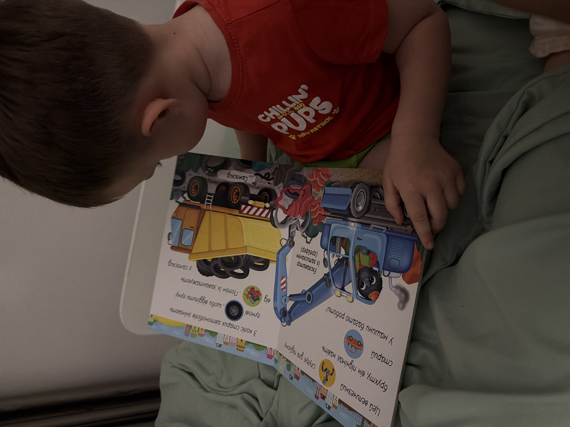 The image shows a young boy in a red t-shirt with "CHILLIN' PUPS" and "PAW PATROL" text, sitting on a light green blanket and looking at an open children's book. The book features illustrations of construction vehicles, specifically a yellow dump truck and a blue excavator. Text in Ukrainian is visible on the pages. The boy's hair is brown, and he is facing away from the camera. The environment appears to be indoors, likely a bedroom, with the soft blanket suggesting a comfortable setting. There is no indication of the location being in Bila Tserkva, Ukraine, or any other specific city.