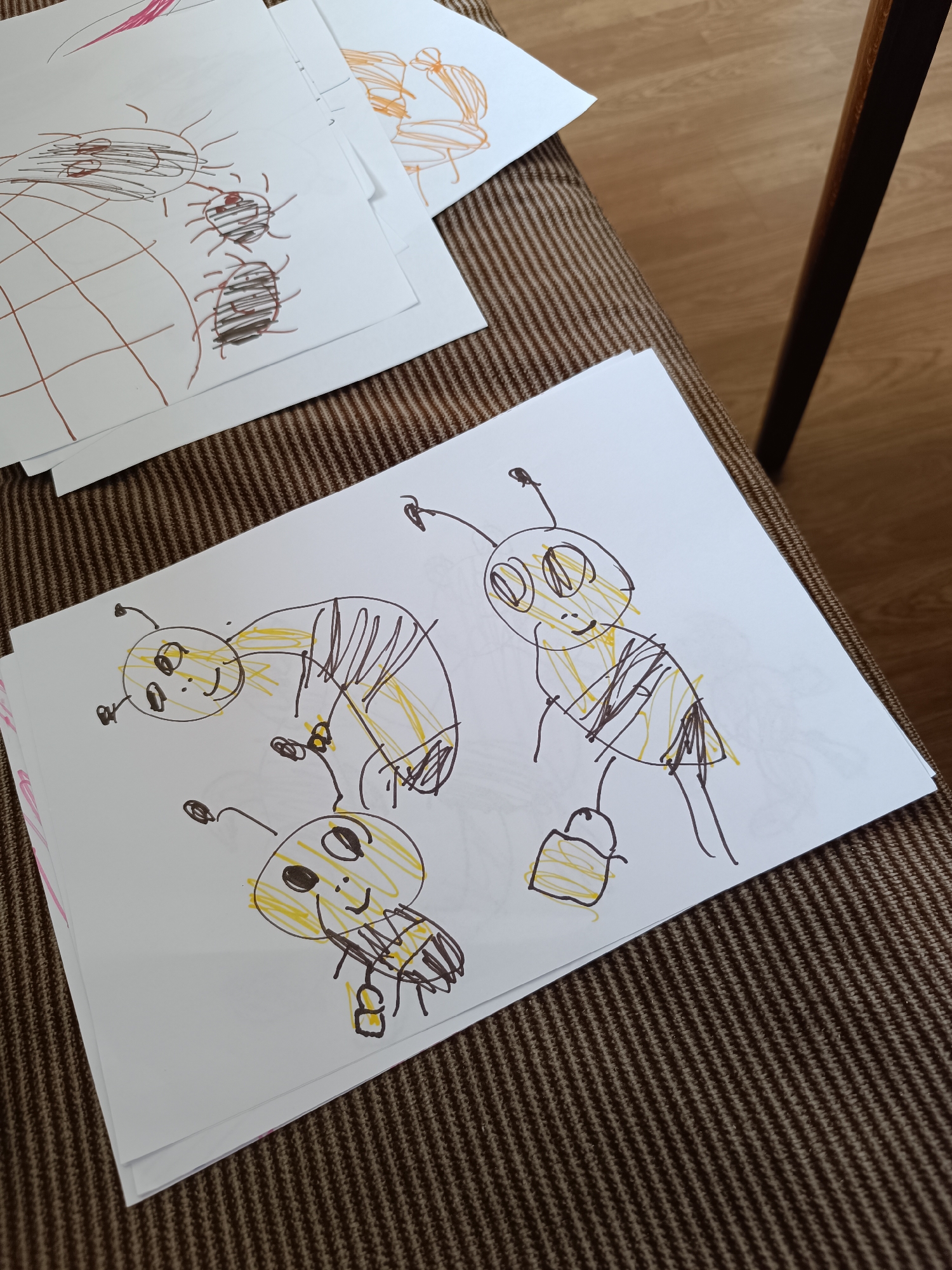 The image depicts a collection of children's drawings on white paper, resting on a brown textured surface. The drawings predominantly feature cartoon-like bees outlined in black and filled with yellow. One drawing shows a grid pattern with spider-like figures. The setting appears to be indoors.