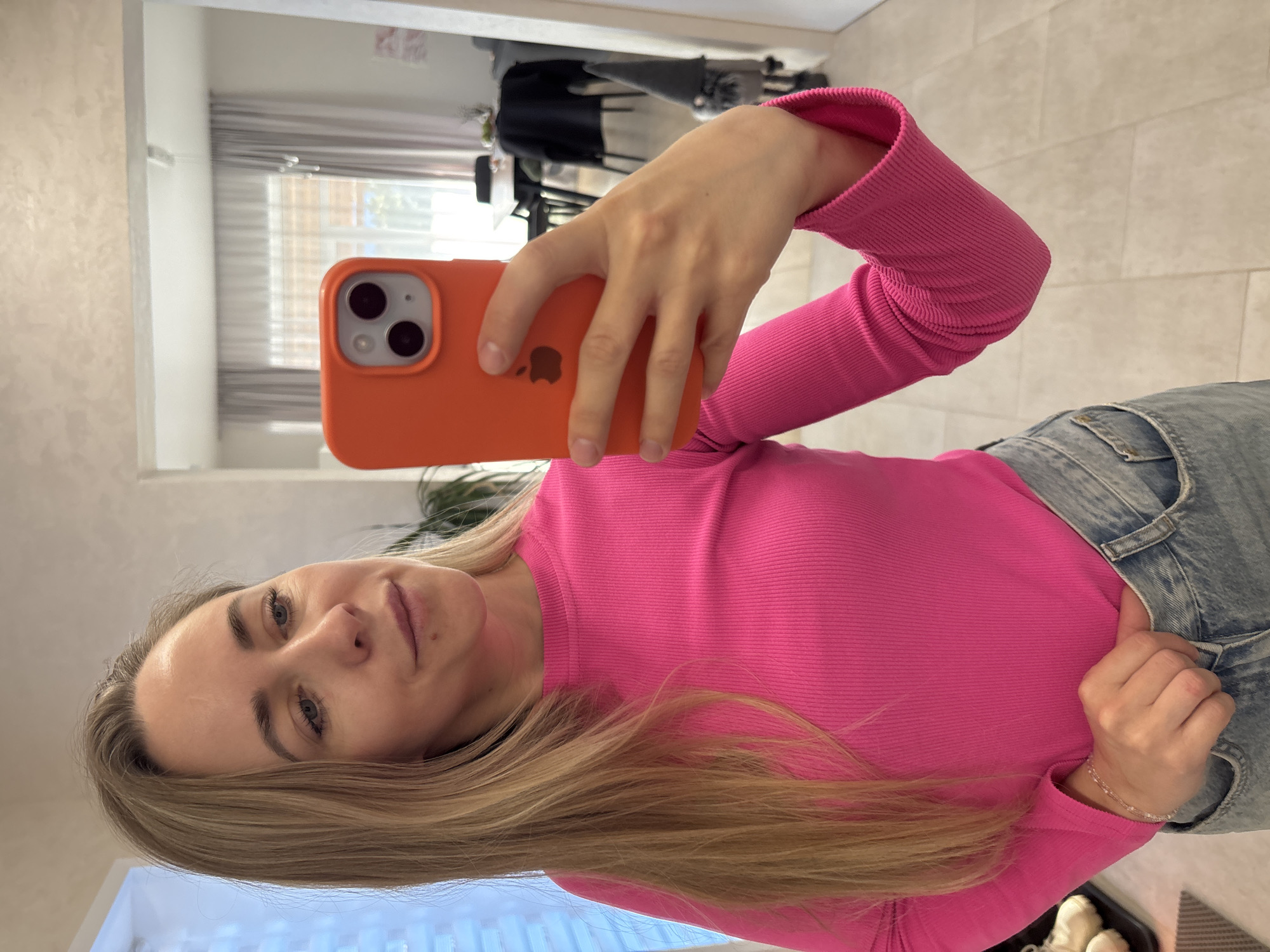 An indoor mirror selfie taken in Chaikovsky, Russia, features a young woman with blonde hair and blue eyes, positioned in what appears to be a brightly lit room. The image is rotated 90 degrees clockwise.
The woman is wearing a vibrant pink, long-sleeved, ribbed top and light blue denim jeans. A delicate bracelet adorns her right wrist. She holds an orange iPhone in a matching orange case, with the Apple logo visible on the back, up to take the photo. Her expression is neutral, looking directly into the camera lens. One hand rests casually on the waistline of her jeans.
The background, reflected in the mirror, shows parts of a room with a light-colored wall or ceiling. A window with horizontal blinds is visible in the reflection, suggesting natural light fills the space. Elements of furniture, possibly chairs or a desk, are also faintly reflected. The floor appears to be tiled. The time of day seems to be daytime due to the natural light.