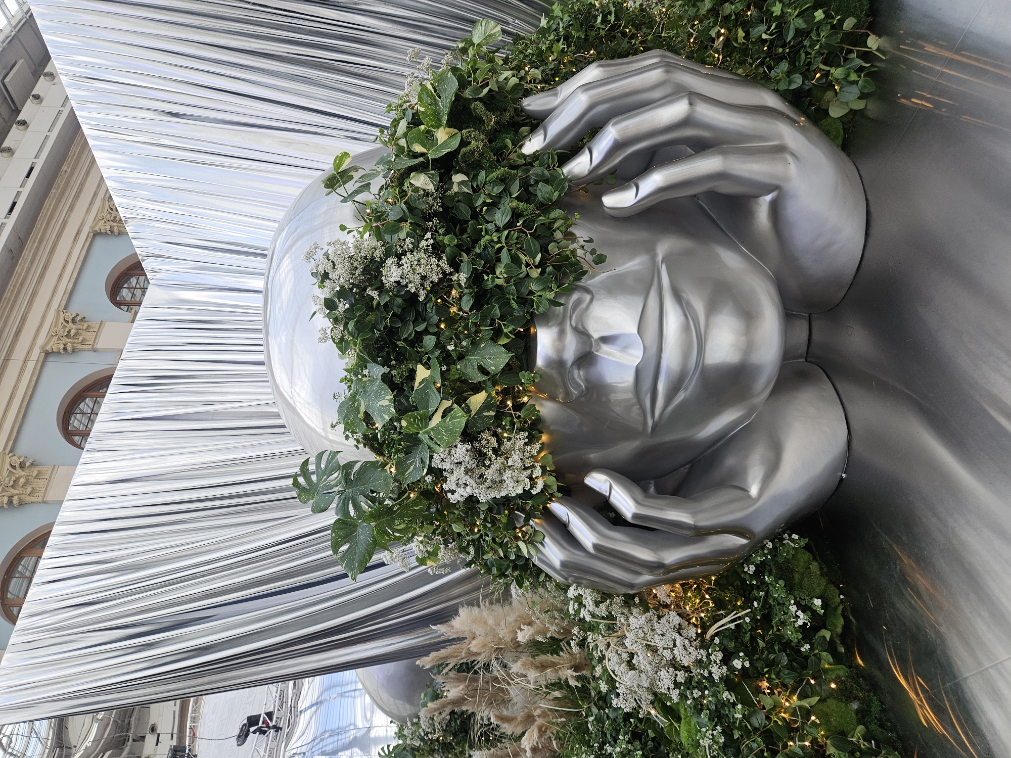 The image shows a silver-colored art installation featuring a human head being cradled by two hands. The sculpture is adorned with lush green vegetation and small white flowers. The installation is set against a backdrop of reflective silver curtains or panels. Through the reflective material, a building with arched windows is visible. This artwork is located in the city of Moscow, Russia. The overall impression is of an artistic presentation within an architectural setting.