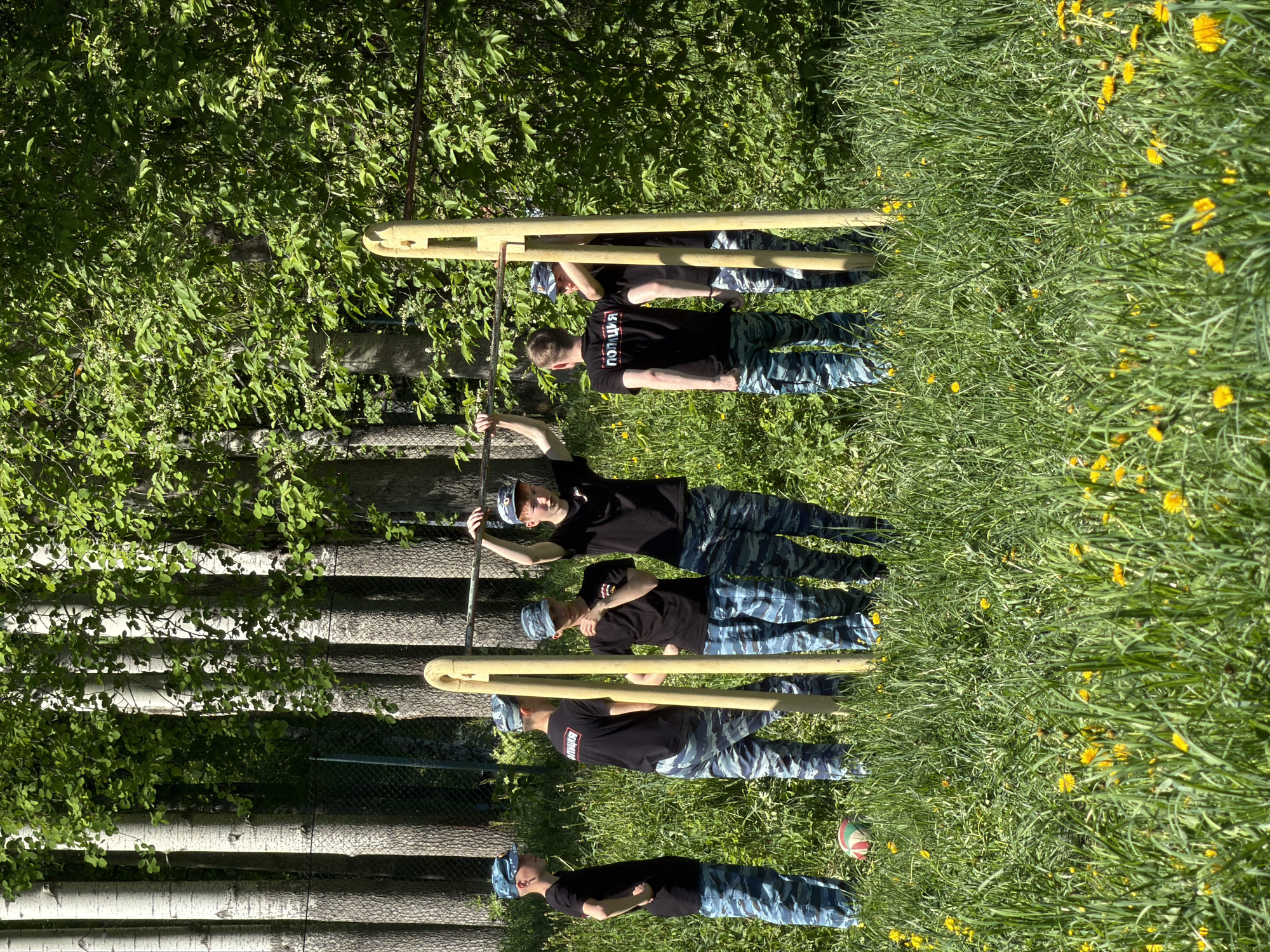 The image shows four individuals wearing black shirts with the word "ПОЛИЦИЯ" (POLICE) printed on them and camouflage pants. They are standing in a grassy area with dandelions. The individuals appear to be performing exercises on a metal structure, possibly a fitness station in a park or training ground. The background includes trees and foliage, and a birch fence. A volleyball is on the ground. It is daytime, with bright sunlight illuminating the scene.
While the description provides a visual account of the scene, it's impossible to pinpoint the exact location within the city of Oreshniki based solely on the image.