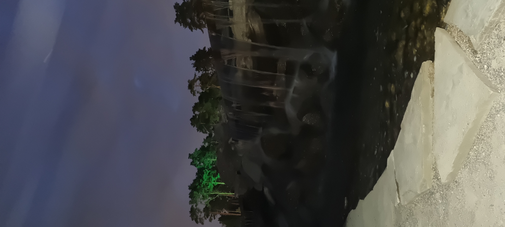 This image depicts a nighttime scene at a park in Shulgino, Russia. A waterfall cascades over dark, rocky terrain, with its water appearing soft and blurred due to a long exposure. The water collects in a dark pool at the base. In the background, a line of trees is visible, with some of them illuminated by green lights, creating a striking contrast against the twilight sky, which is filled with clouds. The foreground features large, light-colored stone slabs placed on a gravel surface, bordering the water.