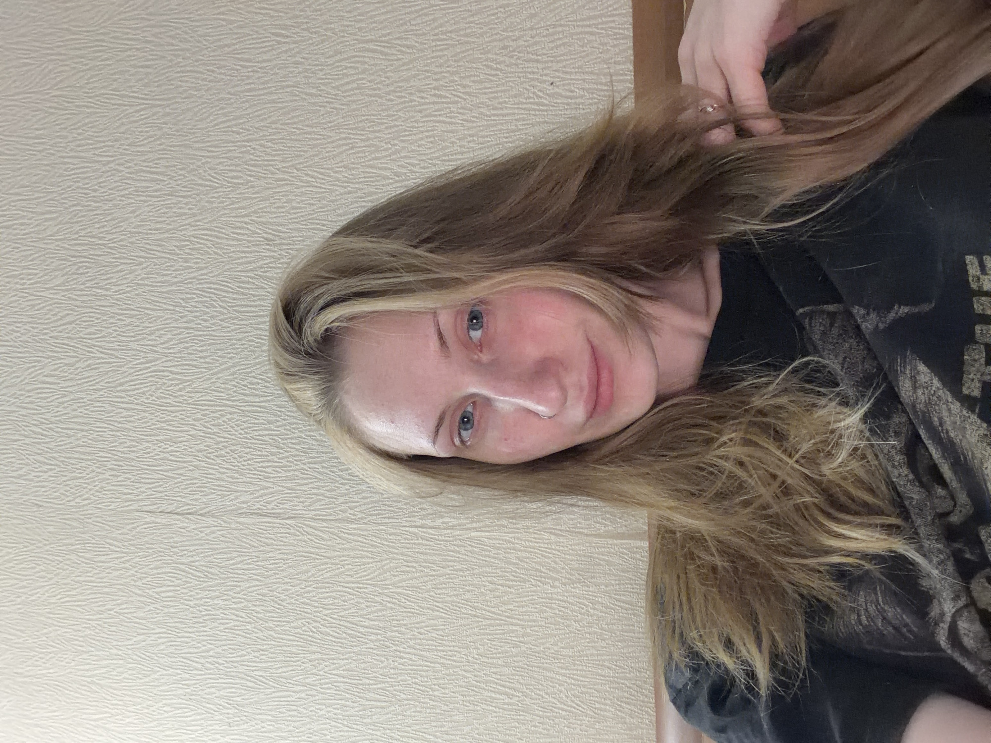 This is a portrait of a young woman with blonde hair, facing the camera. Her hair is long and appears to be slightly wavy, falling over her shoulders and chest. She has light-colored eyes and a fair complexion with some redness on her cheeks. She is wearing a dark t-shirt with a textured pattern. Her left hand is visible near her hair, and she is wearing a ring on one of her fingers. The background is a textured wall, light in color. The lighting suggests an indoor setting, possibly during the daytime. The woman has a subtle smile, conveying a relaxed and natural expression. No visible text is present in the image.