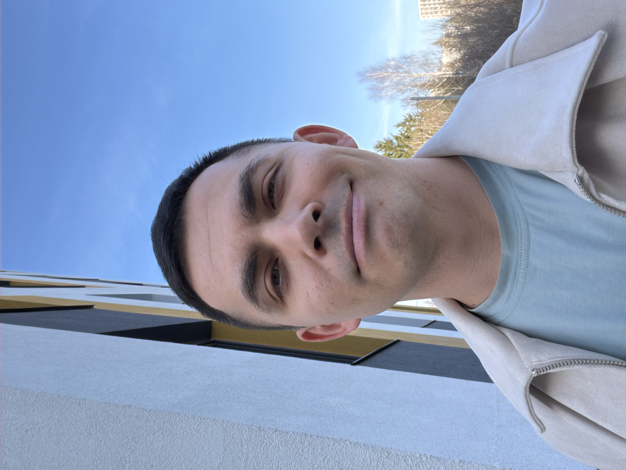 The image displays a single adult male, estimated to be between 25 and 35 years old, in a close-up, direct-to-camera shot. He has short, dark hair, a fair complexion with minimal stubble, and dark eyes, exhibiting a slight smile. He is wearing a light beige collared jacket with a visible silver zipper, over a light blue crew-neck t-shirt. A silver chain is discernible around his neck.
The background on the left features a modern building facade constructed with textured white stucco, dark gray panels, and vertical yellow/gold stripes. To the right, a clear, bright blue sky is visible, with a distant line of trees—comprising both bare deciduous and green coniferous species—and a lighter-colored multi-story building further in the background. The scene is outdoors under natural daylight in Izhevsk, Russia.