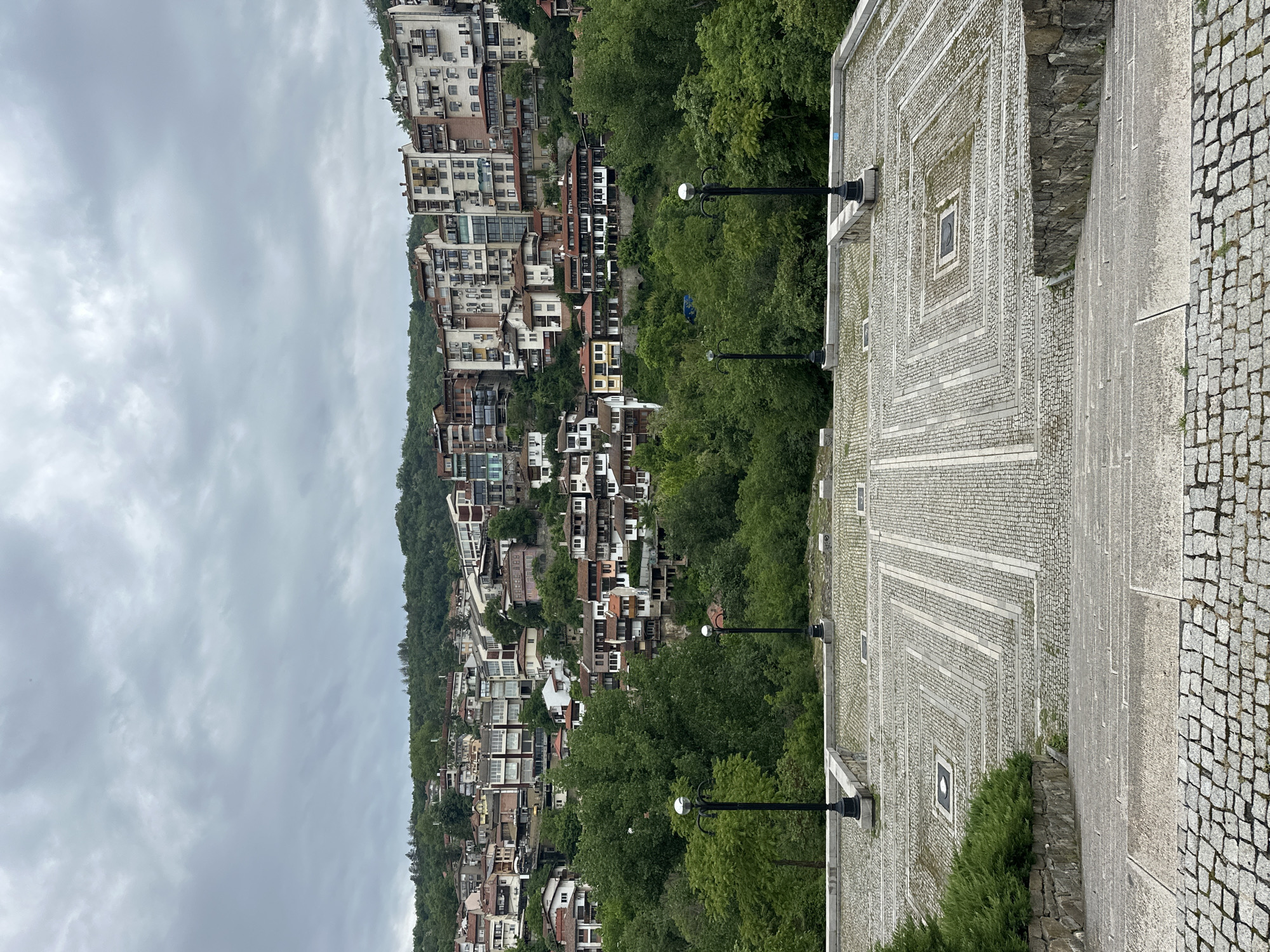 The image captures a view in Veliko Tarnovo, Bulgaria, characterized by a cityscape built on a hillside. In the foreground is a paved area with a geometric pattern consisting of squares within squares, adorned with street lamps. Buildings densely populate the hillside, interspersed with trees. Overcast skies contribute to a muted, somewhat gray atmosphere.