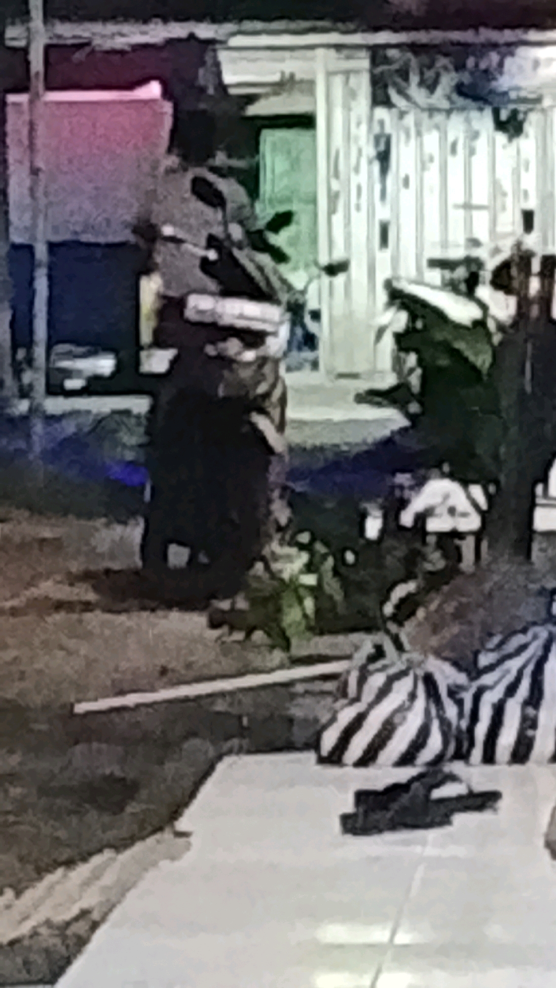 In the city of Ujungrusi, Indonesia, the image depicts a dimly lit, outdoor scene near a structure that appears to be a shop or building. The environment is cluttered with various objects, suggesting a storage area or an informal marketplace. In the foreground, a white surface, possibly a table or a makeshift display, is visible. On this surface, a pair of black sandals are placed. Behind the sandals, two large bags with black and white stripes are situated.
The mid-ground of the image shows several individuals. One person, dressed in dark clothing, is standing and appears to be interacting with a motorcycle. Other figures are partially obscured by objects and shadows, making their details unclear. A motorcycle is visible, positioned to the left of the frame, with its front wheel and handlebars apparent.
The background reveals the facade of a building with vertical elements, possibly shutters or doors. The area behind the building is dark, with hints of what might be foliage or more structures. The overall lighting is low, with some areas illuminated by artificial light sources, casting distinct shadows and adding to the scene's somewhat chaotic appearance. The image quality is low, with a grainy texture and blurred details, making precise identification of all elements challenging.
