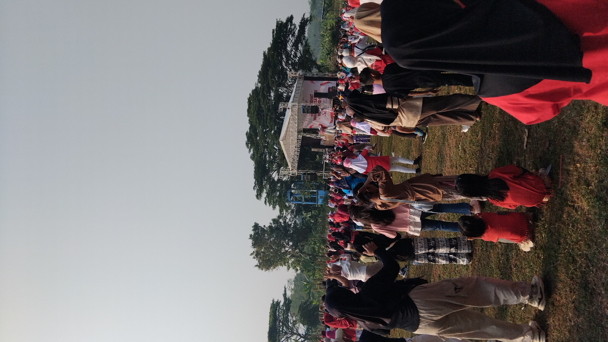 The image shows a large outdoor gathering of people, likely at a public event, with a stage set up in the background. The sky is overcast and bright, suggesting daytime. In the foreground, a group of people, mostly women and children, are spread across a grassy field. Many are wearing headscarves, and the attire suggests a cultural context common in Indonesia. Some individuals appear to be engaged in an activity, possibly exercise or a communal gathering, as some are raising their hands or moving their bodies.
The stage in the middle ground is set up with a white screen or banner displaying text in red and white, partially obscured by the crowd. There are speakers and lighting equipment visible around the stage, indicating a performance or presentation is likely taking place. Behind the stage, trees and a blue structure are visible, suggesting the event is taking place in an open area, possibly a park or sports field on the outskirts of a town. The overall atmosphere seems to be one of a lively community event. The provided context states the location is the city of Posong, Indonesia.