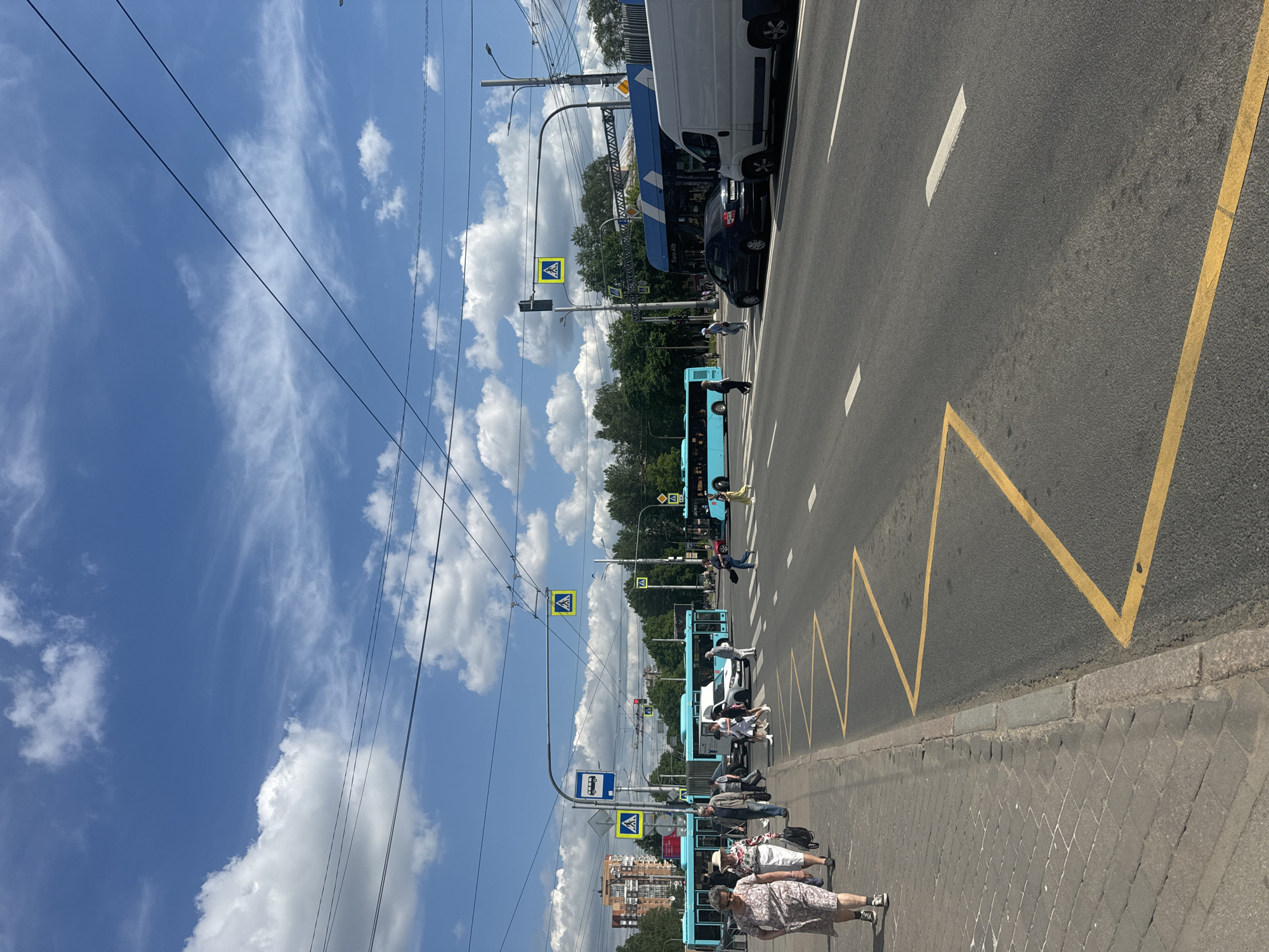 The image captures a street scene in Saint Petersburg, Russia, on a sunny day with scattered clouds. The foreground shows a pedestrian walkway on the left, paved with cobblestones, bordering a street with clear yellow zigzag markings indicating a no-parking zone or a bus stop area. Several people are walking on the sidewalk, including a woman in a floral dress and a straw hat, and a man in a light-colored jacket.
The street itself is a multi-lane road with a pedestrian crossing. Several vehicles are visible: two bright blue buses, one dark car, a white van, and a white car. Pedestrians are crossing the street at the designated crosswalks. Traffic lights and street signs, including a pedestrian crossing sign with a yellow background and a white pedestrian icon, are prominent features of the urban environment. In the background, there are trees and apartment buildings. Overhead, electrical wires for the trolleybuses crisscross the sky. The overall atmosphere is that of a typical urban day with active traffic and pedestrian movement.