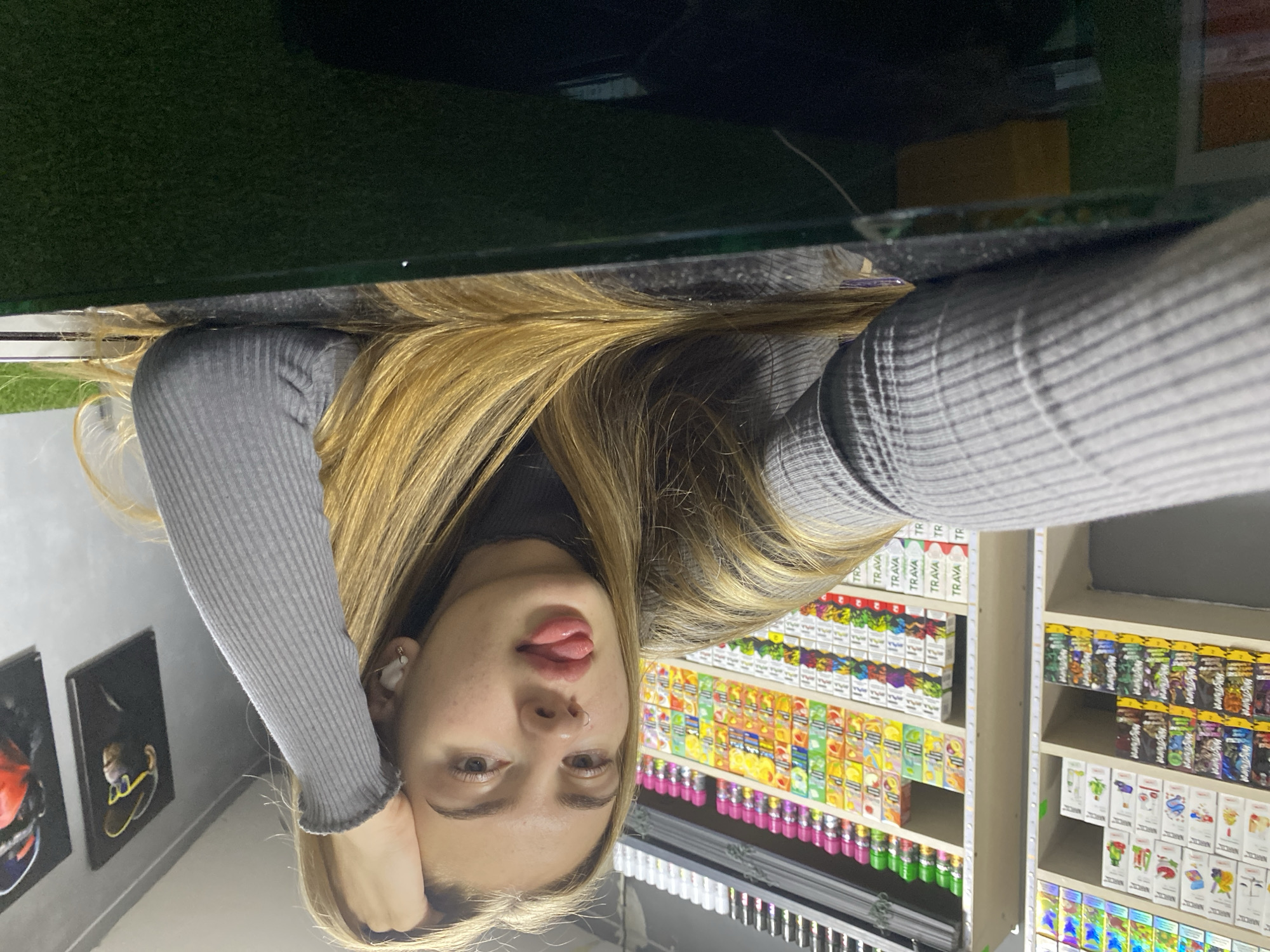A young woman with blonde hair and fair skin takes a selfie. She is wearing a grey ribbed sweater and has her left hand resting on her cheek, her head tilted slightly. Her tongue is sticking out between her lips, and she has a nose ring. In the background, shelves are filled with colorful boxes, possibly vape products or e-liquids. The shelves are brightly lit, suggesting an indoor setting, likely a store. To the left of the woman, two framed pictures hang on a white wall. The overall scene appears to be in a retail environment. The image is taken from a low angle, with the top of the frame showing a dark surface and a glimpse of green material, perhaps artificial turf. The lighting suggests it could be daytime, but the indoor setting makes it difficult to determine. There are no discernible weather cues. Visible text includes "TRAVA" repeatedly on some of the boxes. The location context provided is Дьяково, Russia. The woman's expression appears playful or mischievous.