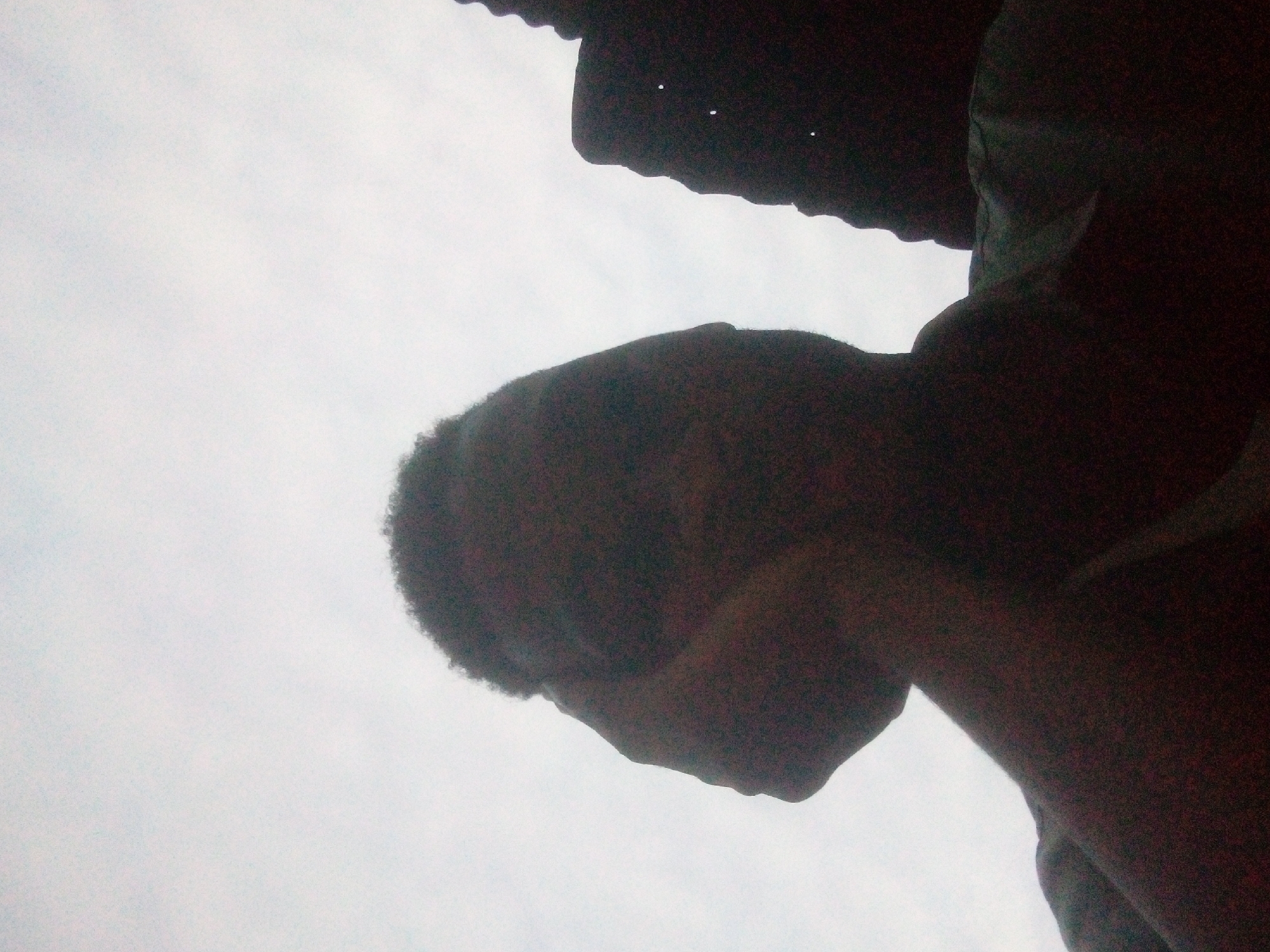 A low-angle shot features a dark-skinned person, likely male, silhouetted against a bright, overcast sky. A roof or overhang is visible at the very top edge of the frame. The location and city cannot be determined from the available information.