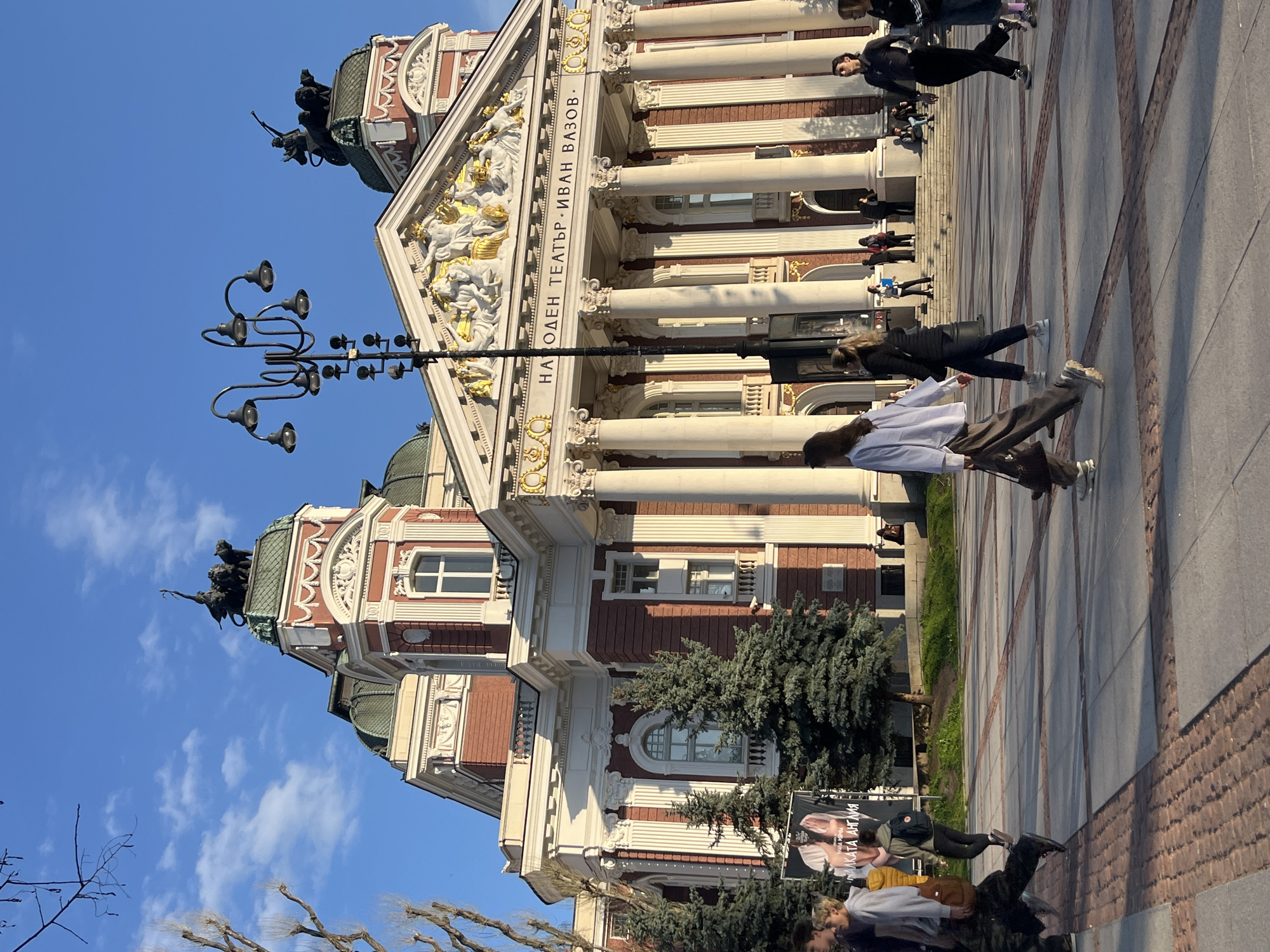 The image captures the Ivan Vazov National Theatre in Sofia, Bulgaria. The building features a grand facade with classical columns, ornate sculptures, and a prominent inscription in Cyrillic. The sky is clear and blue. Several people are walking on the paved plaza in front of the theatre. There is also a vintage street lamp on the left side of the image. A poster is visible near a small tree in front of the theater.