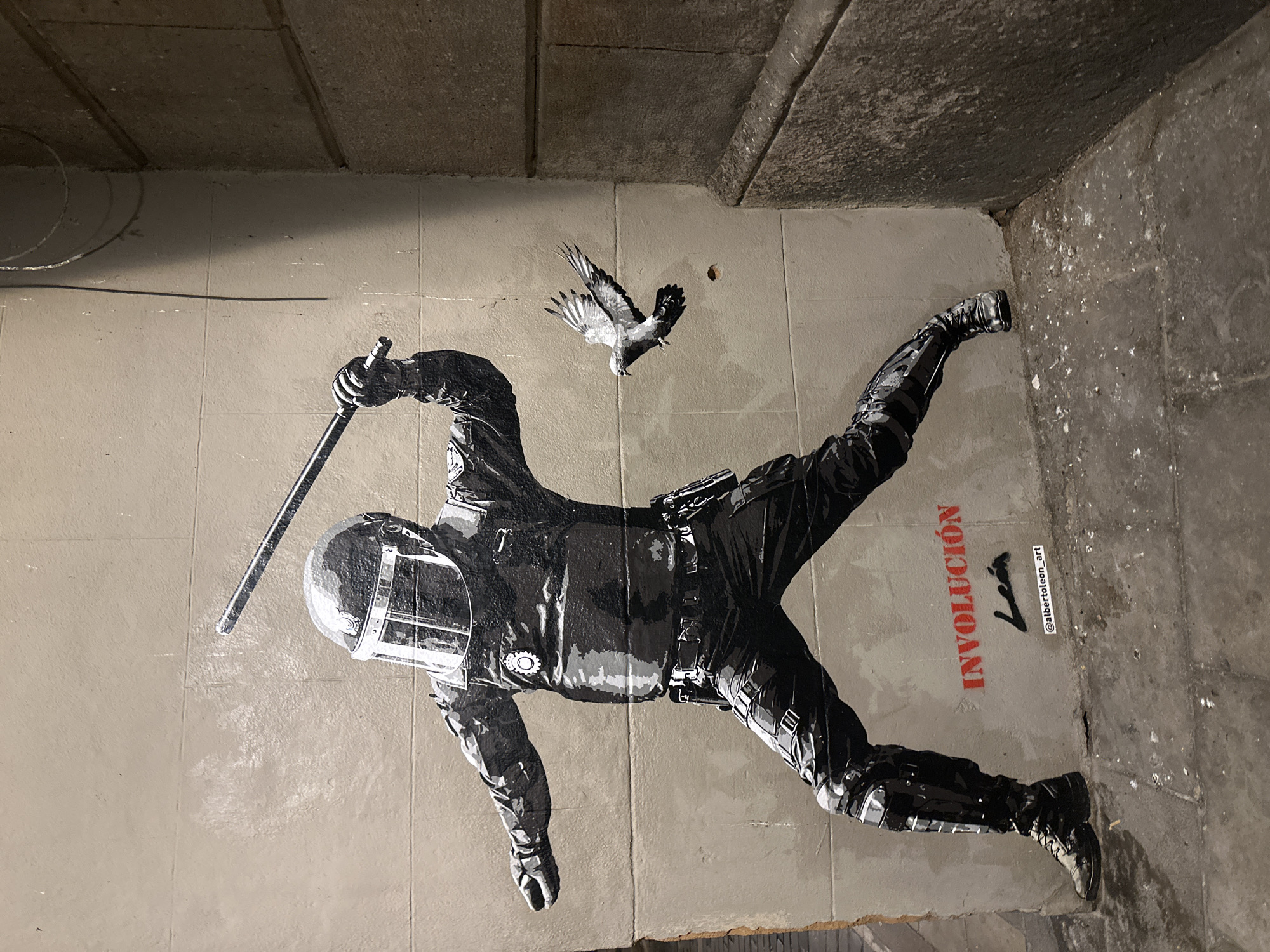 This is a photograph of a mural painted on a concrete wall in a narrow alleyway in Barcelona, Spain. The mural depicts a riot police officer in full protective gear, including a helmet with a visor and body armor, brandishing a baton. The officer is in a dynamic pose, with one leg forward and the other back, as if charging. To the right of the officer, a pigeon is captured in mid-flight, wings outstretched. Below the mural, the word "INVOLUCIÓN" is painted in red, followed by a signature and the Instagram handle "@albertoleon_art". The wall on the left of the mural is smooth and painted a light beige, while the wall on the right is rougher, made of concrete blocks, and appears to be the corner of a building. The ground in the alleyway is dirty and scattered with debris. The overall scene suggests a commentary on social unrest and the role of law enforcement.