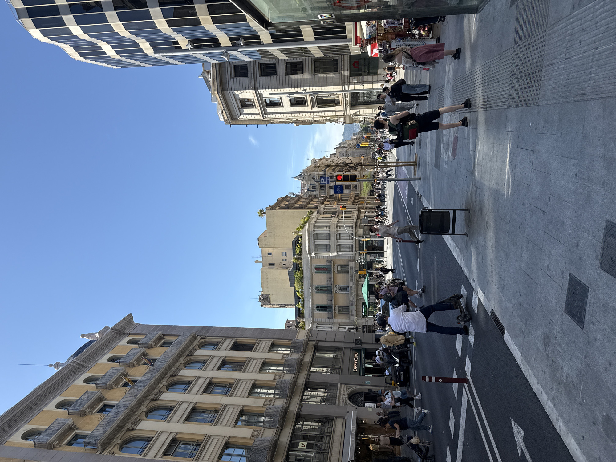 An eye-level, upright shot captures a vibrant urban street scene in Barcelona, Spain, under a clear, bright blue sky on a sunny day. The strong sunlight casts distinct shadows across the pavement.
The wide asphalt street features clear white lane markings and arrows, including a dedicated bike lane on the left side, where several bicycles are visible, some parked, some in use. Pedestrians populate both wide sidewalks, walking, standing, and interacting. In the foreground, a man in a white shirt stands next to an electric scooter, while numerous other individuals, some carrying bags or backpacks, move along the street and sidewalks.
Architectural styles vary significantly. To the left, a tall, classical building with multiple stories, arched windows, and balconies forms the lower part of the frame. On the right, a striking modern building with a black and white checkerboard facade curves overhead, contrasting with the more traditional multi-story buildings further down the street. Some buildings exhibit green foliage on their rooftops, adding a touch of nature. A red traffic light is visible further down the street, and a prominent spire or antenna rises into the sky behind a distant building.
Visible text includes "CAPPICCINO" and "CAFE" on building signs. The overall scene depicts a bustling city environment, full of movement and diverse urban elements.