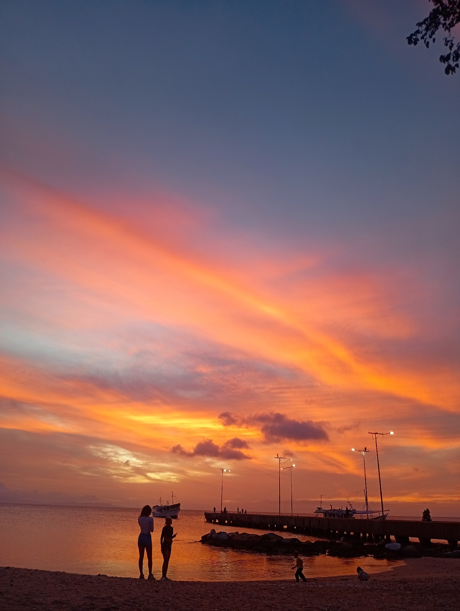The image depicts a sunset scene over a coastal area in Juan Griego, Venezuela. The sky displays a vibrant gradient, transitioning from deep blue at the zenith to shades of purple, pink, orange, and golden yellow near the horizon, with streaks of clouds reflecting these colors. The calm body of water reflects the sky's fiery hues.
In the foreground, a sandy beach is visible, where two adult females stand silhouetted, facing the water. The female on the right holds a rectangular object, likely a mobile phone. Further right on the beach, two smaller figures, identified as children, are present; one is walking towards the left, and the other is crouched on the sand.
A concrete pier extends diagonally from the mid-right into the water, supported by pilings. Multiple individuals are visible as dark silhouettes on the pier, including two figures appearing to ride a motorbike or scooter. Several streetlights along the pier are illuminated. A cluster of rocks forms a breakwater along the pier's base.
In the water near the pier, a larger vessel, possibly a fishing boat or small cargo ship, is anchored. A smaller, less defined boat is present further in the distance to the left of the larger vessel. The upper right corner of the frame shows the dark silhouette of tree branches and foliage.
