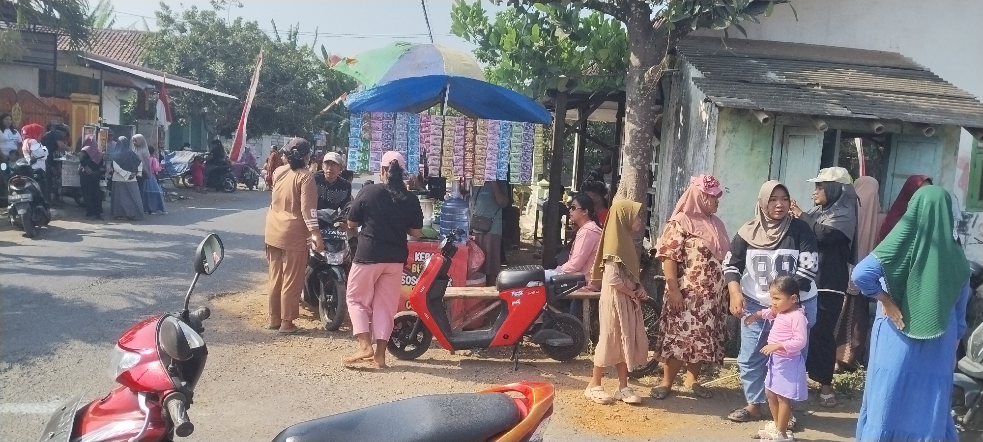In Yosowilangun Kidul, Indonesia, a street scene unfolds under a bright sun. A red scooter is parked in the foreground. To the right, a person wearing a green hijab stands with their back to the camera, their right hand on their hip. In the center, a group of women and children are gathered around a makeshift stall. The stall, adorned with colorful packets of goods and a large umbrella, is situated next to a water dispenser. A red electric scooter is parked at the stall. Several people are interacting, with some standing and others seated. The background reveals buildings with tiled roofs and lush green trees, characteristic of a rural Indonesian setting. Indonesian flags are visible on poles near a building on the left. The overall impression is a lively community gathering at a local roadside vendor.