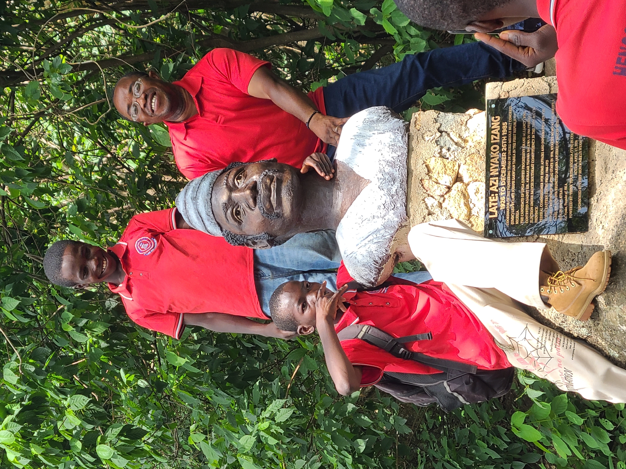 The image depicts a group of three individuals, two young males and one adult male, posing with a bust of a historical figure in an outdoor, natural setting with lush green foliage in the background. The bust appears to be made of bronze and is mounted on a stone pedestal which also features a plaque. The plaque contains text in English, including the name "LATE: AZI NYAKO IZANG" and dates "1926 TO DECEMBER 26TH 1985". The text on the plaque details his role as a coordinator, candidate, and federal commissioner, and mentions his association with "UMBOC", "UPGA", and "Think Tourism Nigeria". The adult male, wearing glasses and a red polo shirt, has his right arm around the bust. One of the young males, also in a red polo shirt and jeans, stands behind the bust smiling. The other young male, in a red polo shirt and khaki pants with a backpack, is positioned to the left of the bust, with his hand near his mouth. The presence of the plaque with Nigerian political and tourism references suggests the location might be a historical site or monument within Nigeria. Based on the text "JOS, NIGERIA" appearing on the plaque as part of the attribution "1926 - 1976 OF THE RIKPEBA BARRA", the location is identified as Jos, Nigeria.