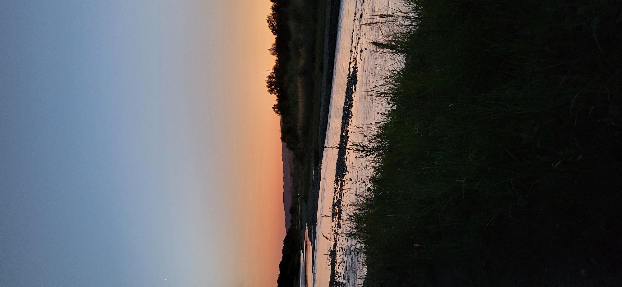 The image depicts a serene natural landscape at dusk, featuring a wide, calm river flowing through a valley. The sky transitions from a deep blue at the top to a warm orange and pink gradient near the horizon, indicating the setting sun. Distant hills and a line of trees form a silhouette against the colorful sky. The river's surface reflects the colors of the sunset and the surrounding environment. The foreground is dominated by lush, dark green grass and reeds along the riverbank, with some pebbles visible in the shallow water. There are no people or animals visible in the frame. The overall atmosphere is tranquil and peaceful.
While the image displays a natural setting, it is not possible to definitively identify the specific location within Taldykorgan, Kazakhstan, based solely on the visual information provided. The scene is consistent with many natural river valleys that could be found in or around the city, but without any identifying landmarks or textual context, a precise location cannot be determined.
