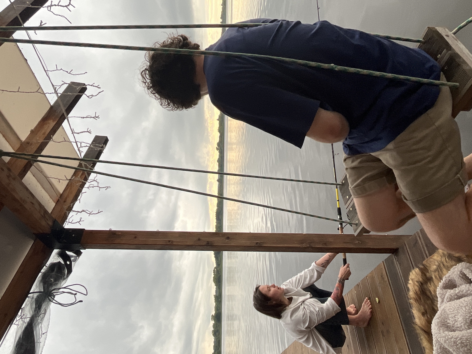 The image captures a serene evening scene by a body of water, possibly a lake or river, with a hint of sunset on the horizon. The location is identified as Radchenko, Russia. Two individuals are present, engaged in fishing.
On the left, a person is crouched on a wooden dock, wearing a light-colored long-sleeved shirt and dark pants. They are actively holding a fishing rod, with tattoos visible on their arm. Their gaze is directed towards the water, indicating focus on their activity. The dock is constructed from wooden planks, and a fuzzy blanket or rug lies nearby.
On the right, another individual is seated on a swing, facing away from the camera and towards the water. They are wearing a dark blue t-shirt and light brown shorts. They are also holding a fishing rod, suggesting they are fishing concurrently. The swing is suspended by ropes from a wooden structure, which also supports a roof or canopy. This structure appears to be an overhang or part of a boathouse.
The environment is characterized by calm, reflective water, a tree-lined distant shore, and an overcast sky with soft, diffused light. String lights are visible hanging from the wooden structure, adding a decorative element to the scene. The overall atmosphere is one of peaceful outdoor recreation.