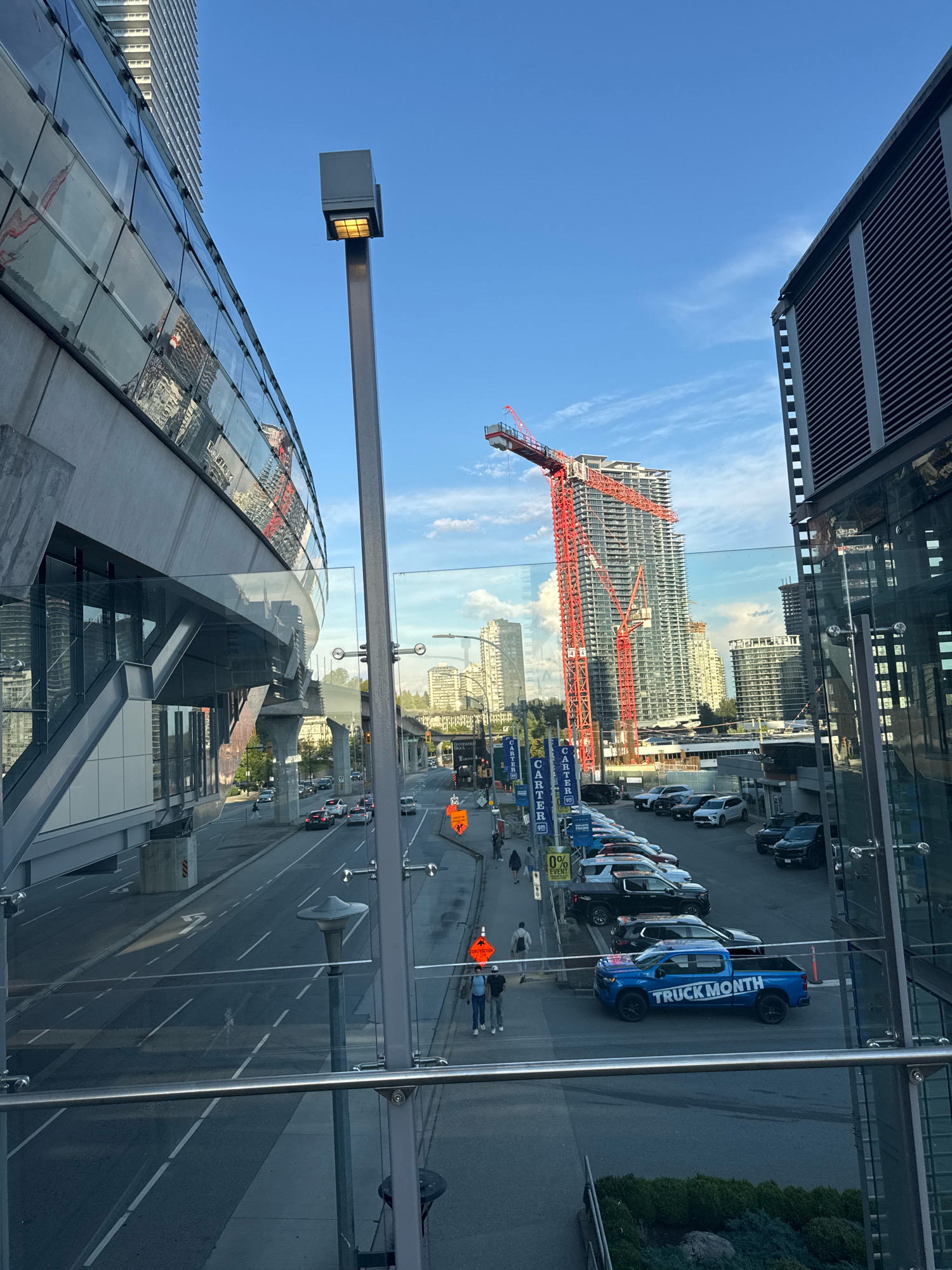 The image depicts an urban street scene in Burnaby, Canada, likely near Brentwood Town Centre, characterized by modern architecture and ongoing construction. In the foreground, a glass railing of an elevated walkway or building offers a view of the street below. A prominent light pole with a rectangular fixture stands near the railing.
To the left, a sleek, modern building with a curved facade of glass panels reflects the surrounding environment, including a tall, white building in the distance. To the right, another contemporary building with louvred vents is visible.
The street below is a multi-lane road with moderate traffic. Several cars are parked along the right side of the street. A bright blue pickup truck with "TRUCK MONTH" emblazoned on its side is parked prominently on the road. Further down the street, a large construction site is active, dominated by a tall, red tower crane extending over a high-rise building under construction. Several smaller buildings and other construction cranes are also visible in the background, indicating significant development.
Pedestrians can be seen walking on the sidewalks and crossing the street. Construction signage, including orange warning signs, is present along the roadside. The sky is a clear blue with a few wispy clouds. The overall impression is of a dynamic urban environment undergoing transformation.