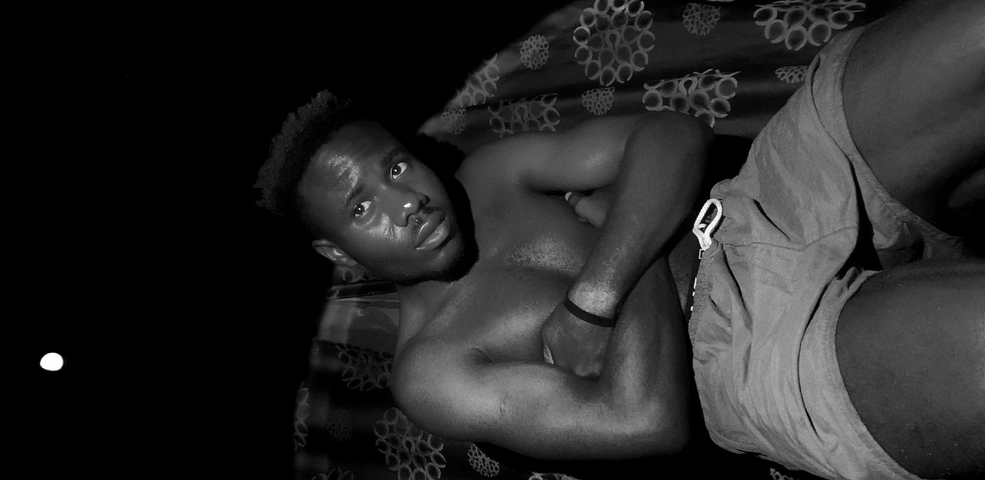 A low-angle, black and white photograph depicts a shirtless black man lying on a surface with a patterned covering. The man's torso is exposed and muscular, and he is wearing shorts. His left arm is bent across his chest, his hand near his shoulder, and he is wearing a dark wristband. He is looking toward the camera. A small, bright circular object is visible in the dark area to the left of the man. The location is said to be Narayi, Nigeria, but the image itself contains no contextual clues to verify this information.