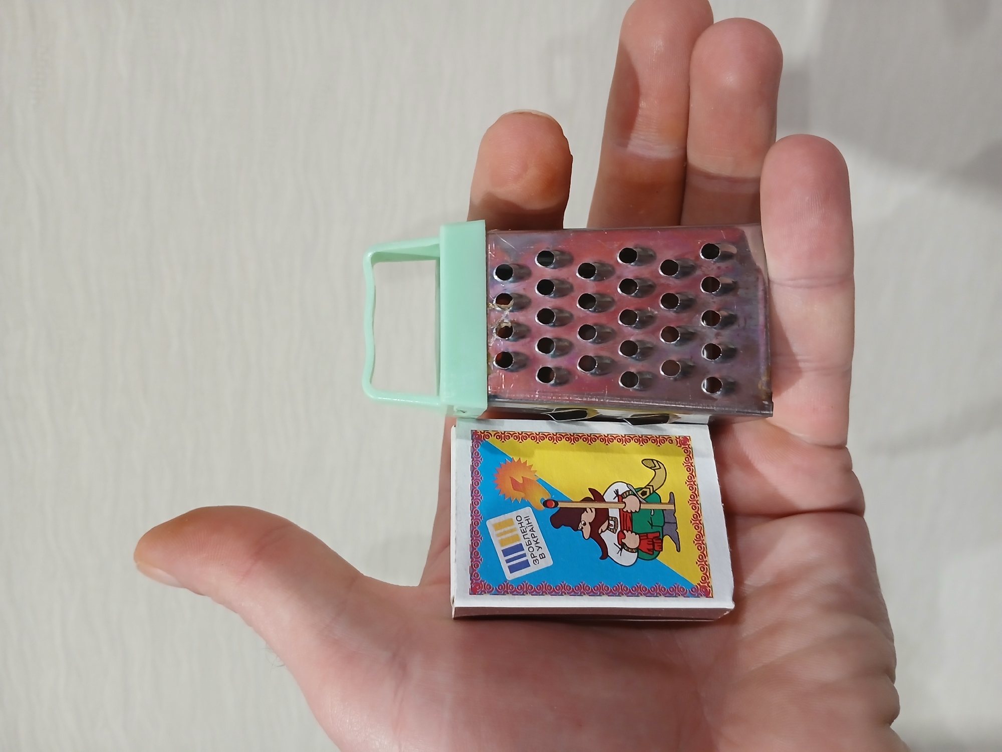 A person's hand holds a small, possibly miniature, cheese grater with a light green plastic handle on top of a box of matches. The box of matches has a cartoon illustration of a Cossack-like figure holding a lit match, with the words "ЗРОБЛЕНО В УКРАЇНІ" (Made in Ukraine) in blue and yellow colors. The grater appears to be made of metal and has numerous holes. The background is a plain, light-colored surface. There is no information available to confirm this scene's exact location or details in Kamianske, Ukraine.