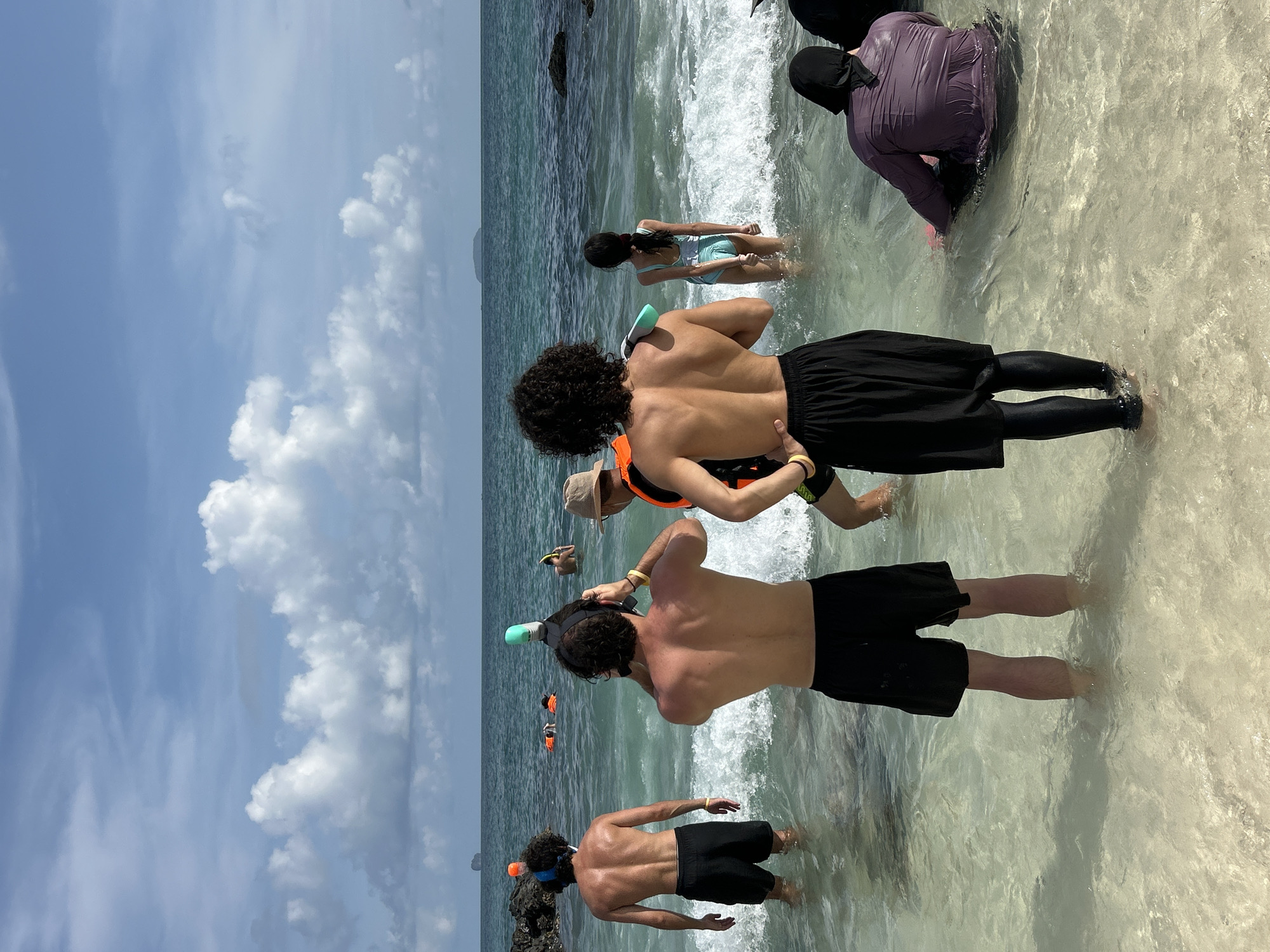 The image depicts a group of people in shallow, clear ocean water, likely near a beach in Phru Nai, Thailand. The sky is a mix of blue and white clouds, with a distant island visible on the horizon. Several individuals are equipped with snorkeling gear, suggesting an activity in progress or about to begin.
In the foreground, two shirtless men are facing away from the camera, with their backs to the viewer. One is wearing black shorts and the other is also in black shorts. They appear to be standing in the water, with waves gently lapping around their legs. Behind them, a person in a tan hat and an orange life vest is partially visible, also facing away from the camera.
Further into the water, another shirtless man with curly hair is visible, wearing black shorts and snorkeling gear. A woman in a light blue swimsuit is also in the water, facing away from the camera. To the far right, two individuals are partially submerged in the water, one wearing a purple, long-sleeved garment and headscarf, and another person in darker clothing.
In the middle ground, several people are in the water, some wearing orange life vests, suggesting a snorkeling tour or group activity. The water is a translucent turquoise color, with a sandy bottom visible in the shallower areas. Rocks are present along the shoreline to the left. The overall scene conveys a relaxed, summery atmosphere associated with a beach destination.