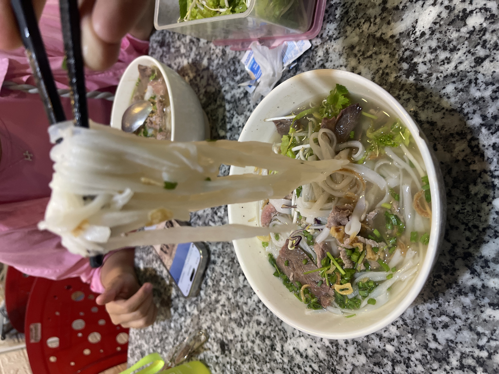 The image presents an overhead view of a casual dining scene, likely in Bac Nha Trang Ward, Vietnam, centered around two bowls of pho. In the foreground, a person's hands, wearing a pink long-sleeved shirt, are actively using black chopsticks to lift a substantial portion of long, flat white rice noodles from a white bowl. The noodles appear translucent and are interspersed with small pieces of green herbs.
The bowl contains a clear broth, numerous thin slices of beef, bean sprouts, chopped green onions, and fresh cilantro or other herbs, indicating a traditional Vietnamese beef noodle soup. Another similar white bowl of pho, with a spoon resting inside it, is partially visible in the upper left background.
To the upper left of the main bowl, a clear plastic container holds a generous amount of fresh green herbs and leafy vegetables, typically served as accompaniments with pho. The table surface is a speckled granite or marble pattern. Other items on the table include a smartphone lying flat, its screen unreadable, and a crumpled small white plastic bag with faint blue text on it. Part of a red plastic stool or chair is visible on the far left, and a green clip or utensil holder is at the very bottom. The lighting suggests an indoor setting, and no specific time of day is discernible. No emotions are visible, and any text on the bag or phone is too blurry to decipher.