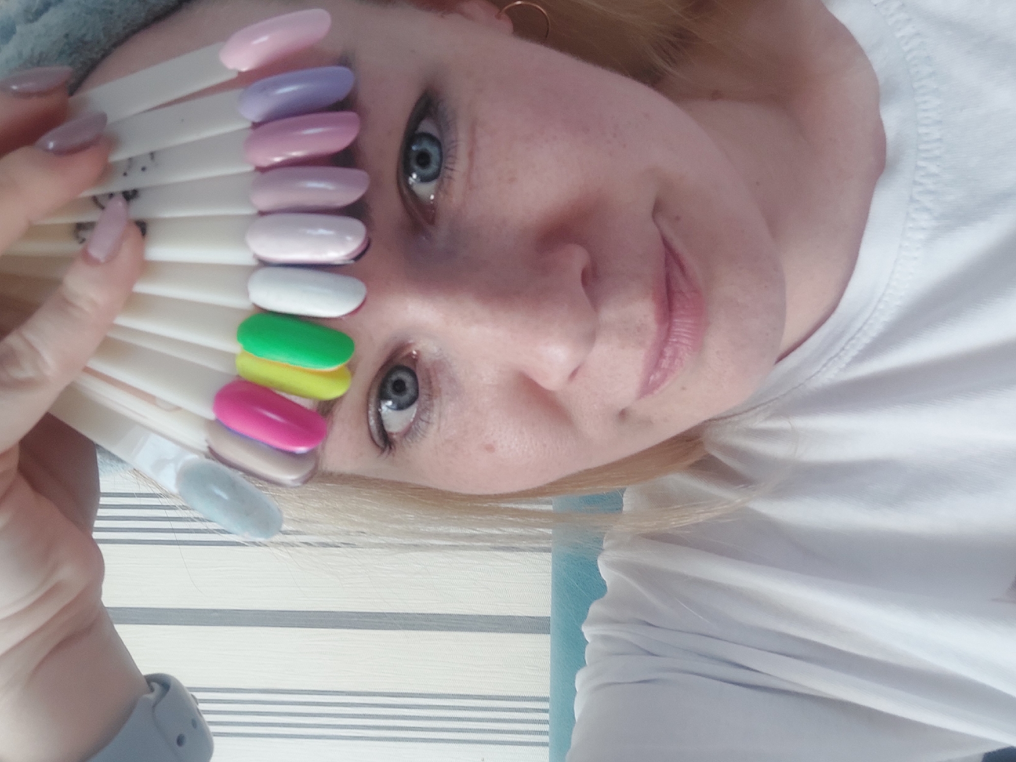 A woman with blonde hair and blue eyes is holding a fan of nail polish color swatches. The colors include light pink, lavender, rose pink, light peach, white, green, yellow-pink, and light blue. She is wearing a white shirt and gold hoop earrings. The background consists of a striped wall. It is not possible to verify if the image was taken in Chaikovsky, Russia, based on the provided information.