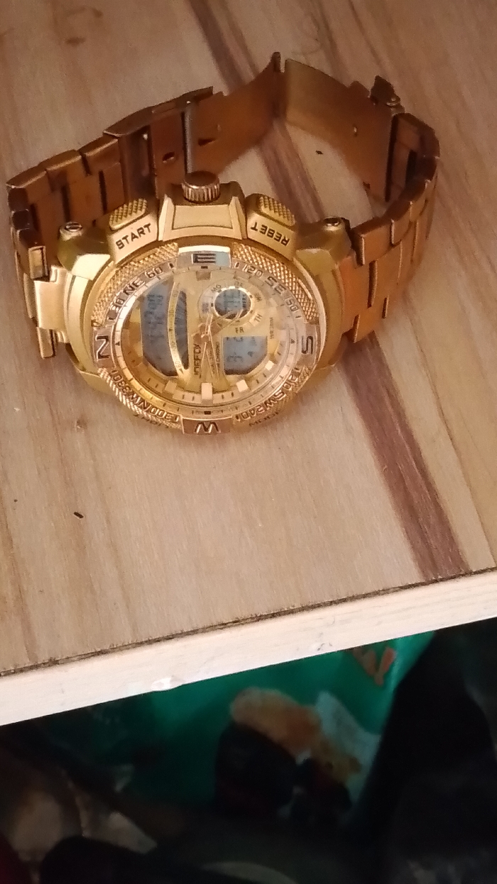 A gold-colored JINOFFLY wristwatch is displayed on a light-toned wooden surface in Ogboko, Nigeria. The watch features a round, multi-functional analog-digital dial with a metallic link band. Visible text on the watch case includes "START" and "RESET," while the bezel displays compass points such as "N," "NE," "E," "SE," "S," "SW," "W," and "NW," alongside numerical indicators. The watch's metal strap is unclasped and partially extended. The wooden surface exhibits a vertical grain pattern. Below the visible edge of the wooden surface, the background is dark and out-of-focus, revealing indistinct multi-colored objects, including a prominent green item and blurred shapes of orange, white, and black.