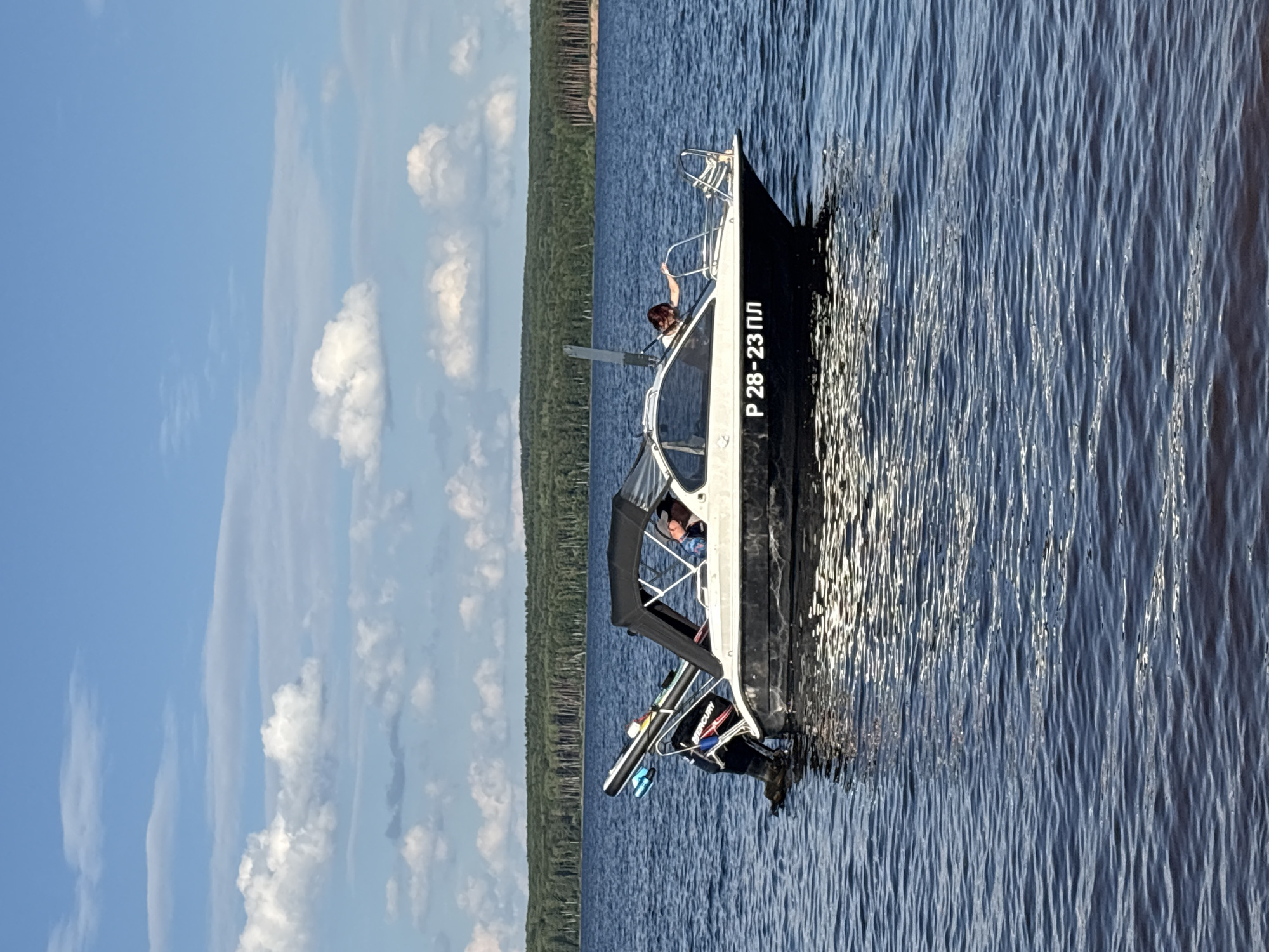 This media file shows a boat on a body of water, likely a lake or large river, with a forested shoreline in the background. The sky is a clear blue with scattered white clouds, suggesting fair weather, and the time of day appears to be daytime, likely afternoon given the lighting. The water is a deep blue and shows ripples from the movement of the boat and possibly a gentle breeze.
The boat is a dark-hulled vessel with a white cabin structure, partially covered by a black canopy. There are at least two people visible on the boat. One person is leaning over the side, seemingly interacting with something outside the boat. Another person is inside the cabin, partially obscured. On the stern of the boat, an outboard motor is visible, which is black with red and white accents.
A notable detail on the side of the boat is visible text in white lettering: "Р 28-23 ПЛ". This is likely a registration number or identifier for the boat. The overall mood is peaceful and recreational, suggesting a day spent out on the water. The presence of the boat and the natural setting point to an outdoor leisure activity. The location context of Chaikovsky, Russia, suggests this scene is set in that region, known for its waterways and natural landscapes.