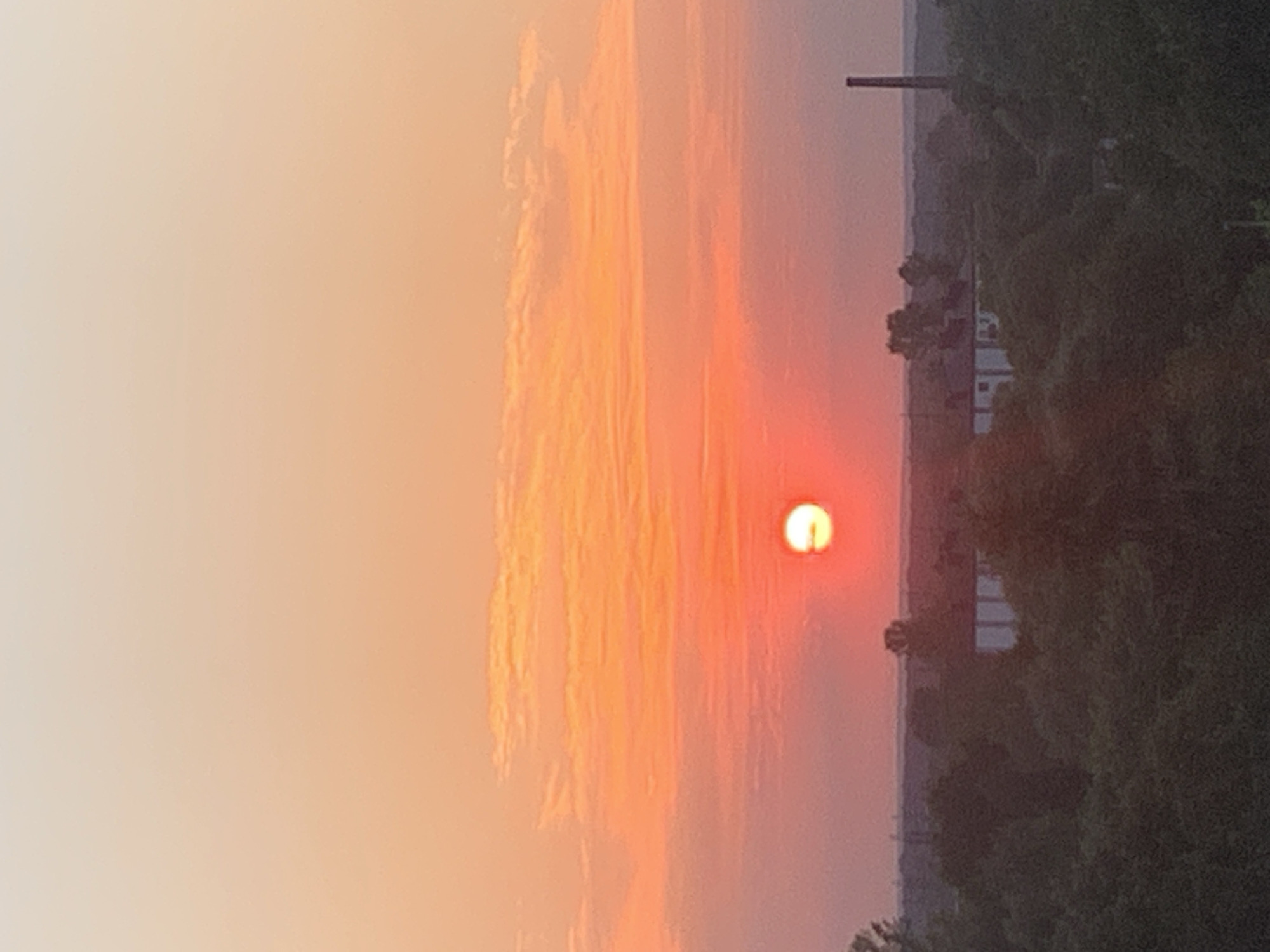 The image shows a sunset over the city of Kolomyia, Ukraine. The sun, a bright orange orb, is partially obscured by clouds in the sky, which transitions from a pale yellow at the top to a vibrant orange and pink near the horizon. Below the sky, a dark silhouette of trees and buildings forms the landscape. In the distance, a tall, slender smokestack rises above the treeline. The overall impression is one of a peaceful, late afternoon in an urban setting.