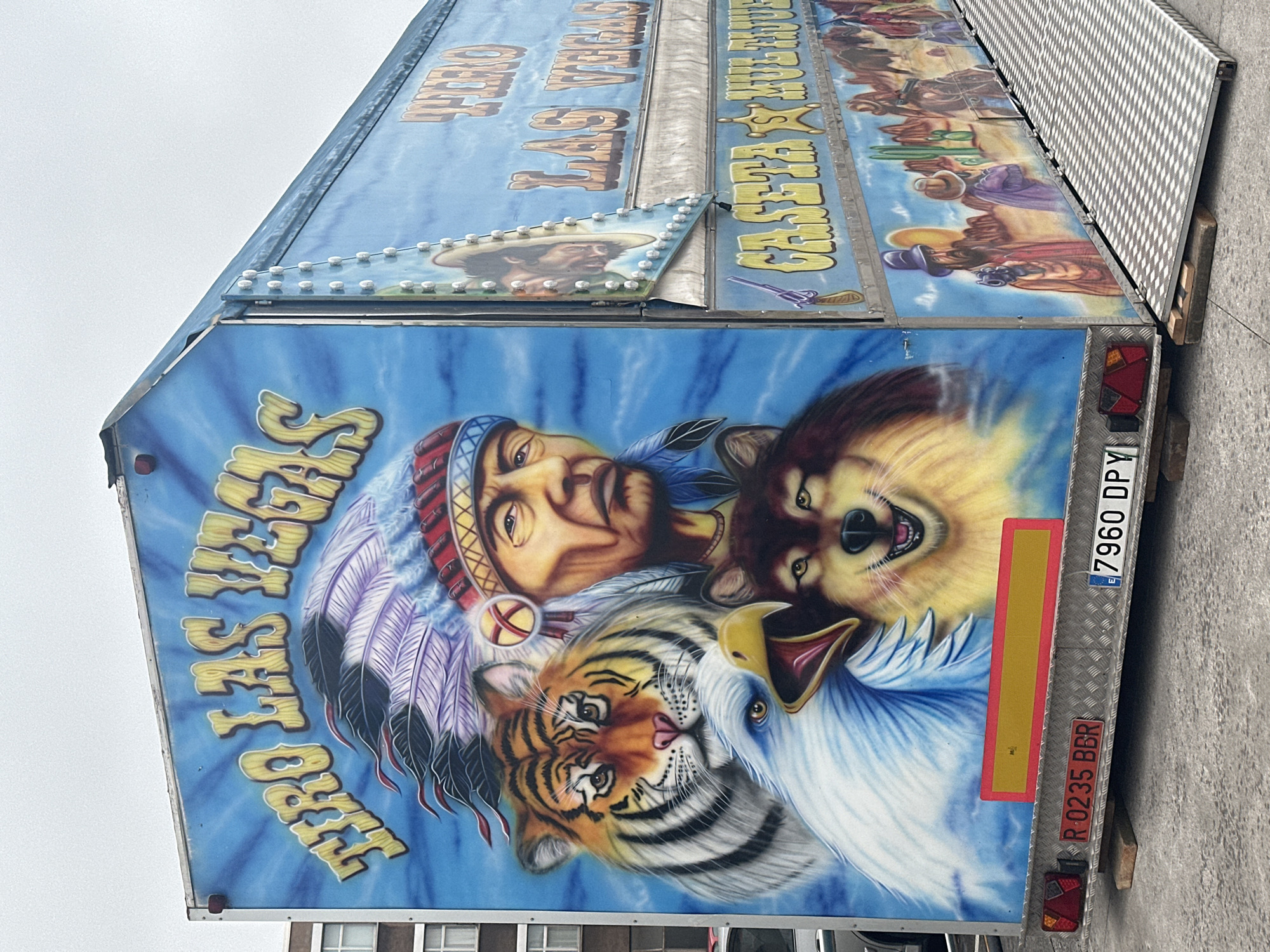 The image shows a colorful trailer with murals depicting a Native American chief, a tiger, an eagle, and a wolf. The text "TIRO LAS VEGAS" is prominently displayed on the side of the trailer. There are also scenes of cowboys and a desert landscape. The trailer appears to be set up for an event, possibly a carnival or fair, as indicated by the ramp leading up to the entrance and the license plates on the rear. The trailer is parked outdoors on a paved surface, with a building visible in the background. The sky is overcast, suggesting a cloudy day. The license plate on the left reads "R 0235 BBR", and the license plate on the right reads "7960 DPY". The trailer is positioned on wooden blocks. It is difficult to ascertain the exact location within the city of Gijón, Spain without further context or distinguishing landmarks.