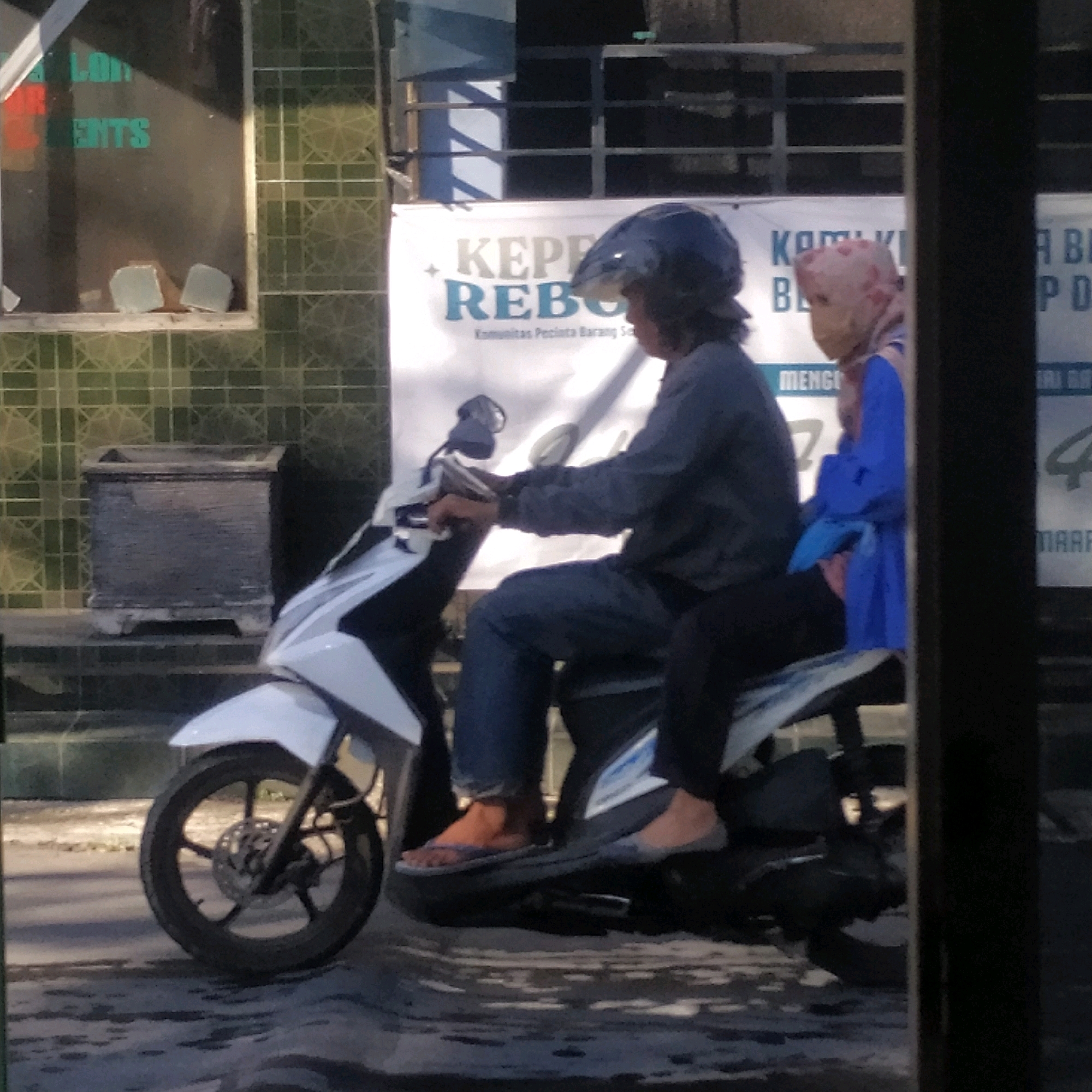 Two individuals are riding a white scooter on a street in Batan, Indonesia. The person in front, wearing a dark blue helmet and a dark grey long-sleeved shirt, is driving. The person behind, wearing a light pink hijab and a light blue garment, is the passenger. The scooter is white with black accents. In the background, there are buildings with tiled walls and a banner with text in Indonesian. To the left of the scooter, there is a large rectangular planter box. The road surface appears wet, with reflections visible. The lighting suggests it is daytime.