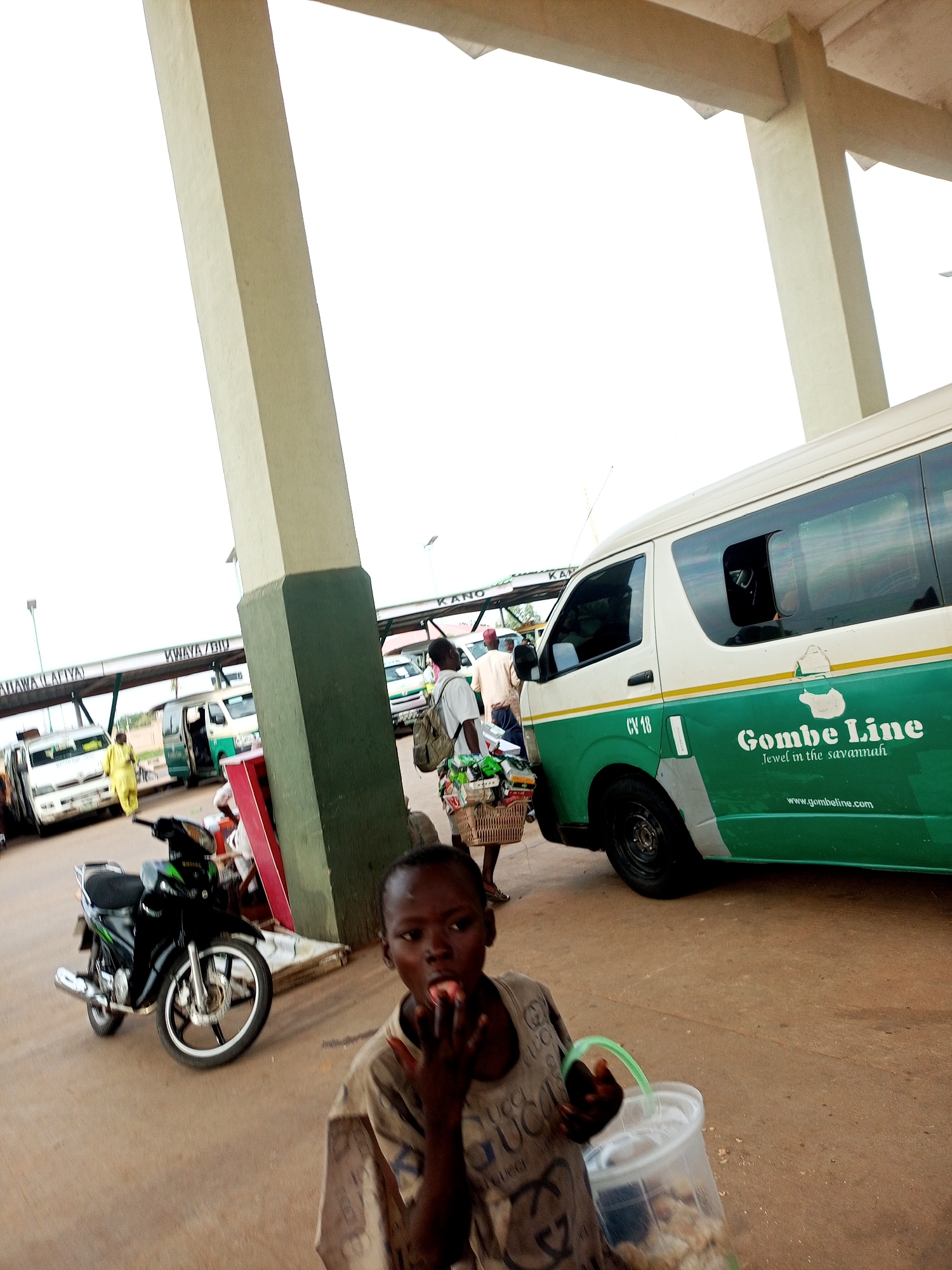 The image captures a scene at a bus station in Gombe, Nigeria. In the foreground, a young boy is seen eating something while holding a plastic container. He is wearing a light-colored t-shirt with a patterned design. Behind him, to the left, is a parked black motorcycle. Further back, various vehicles are visible, including a white and green bus labeled "Gombe Line" with the website "www.gombe line.com" printed on its side. Other vehicles, possibly minibuses and vans, are also present. Several people are dispersed throughout the station, some carrying luggage and baskets, suggesting travel and commerce. The environment appears to be an open-air bus terminal with covered waiting areas supported by pillars. Signs indicating routes like "KANO" and "KWANYA/DIU" are visible on overhead structures, identifying it as a transportation hub. The ground is paved, and the overall lighting suggests it is daytime.