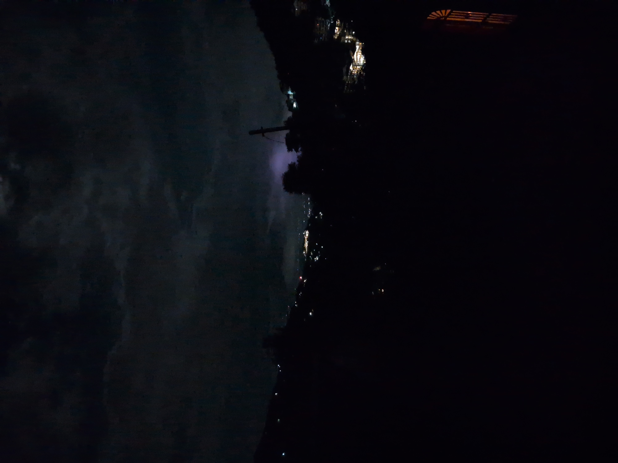 This is a night time, long shot, landscape image of a valley with a city in the distance. The city of Haini, India is visible as a cluster of lights on a hillside in the middle ground. There is a faint purple glow in the sky above the city, possibly from lightning or artificial lighting. A wooden gate or fence is visible in the foreground on the right side of the image. The sky is dark and cloudy, with some of the clouds illuminated by the distant city lights. The overall scene is dark and moody, with the city lights providing the only significant source of illumination.