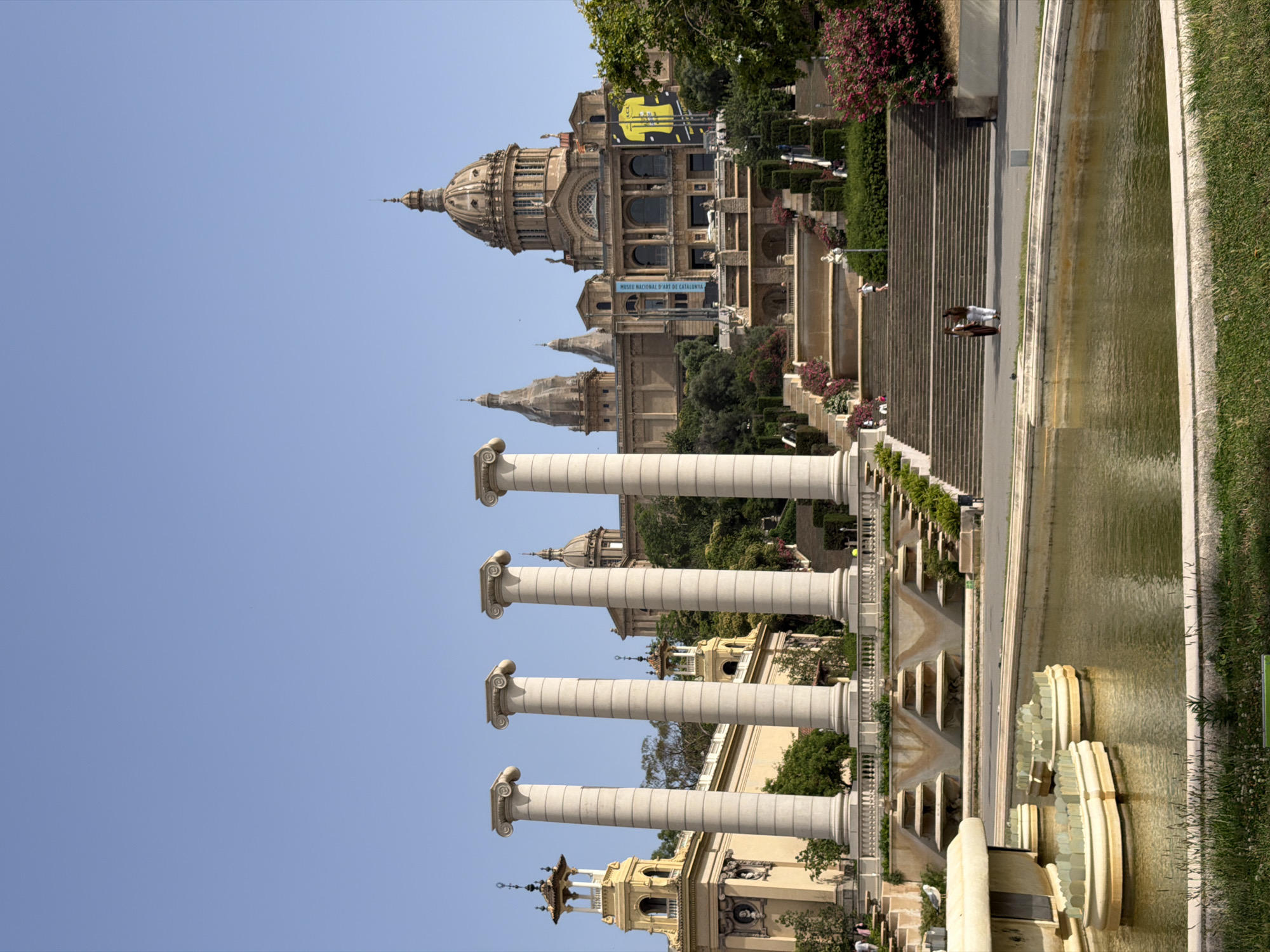 The image depicts the exterior of the Palau Nacional, a grand building in Barcelona, Spain. The building features a prominent dome and classical architectural elements. In the foreground, a series of large, fluted columns stand in a line, suggesting a monumental approach to the structure. A water feature, possibly a fountain or canal, runs alongside a set of stairs leading up to the building. The water appears murky green. On the steps, two individuals are visible, one wearing white and the other in darker clothing. The sky is clear and blue, indicating fair weather. Lush green grass lines the edge of the water feature. Trees and flowering bushes add to the scenic setting. A banner with text that appears to be in Catalan is visible on the building. Another banner, a blue one, also has text on it. The overall scene suggests a daytime visit to a significant cultural landmark. The architecture, landscaping, and presence of visitors imply a tourist or recreational context.