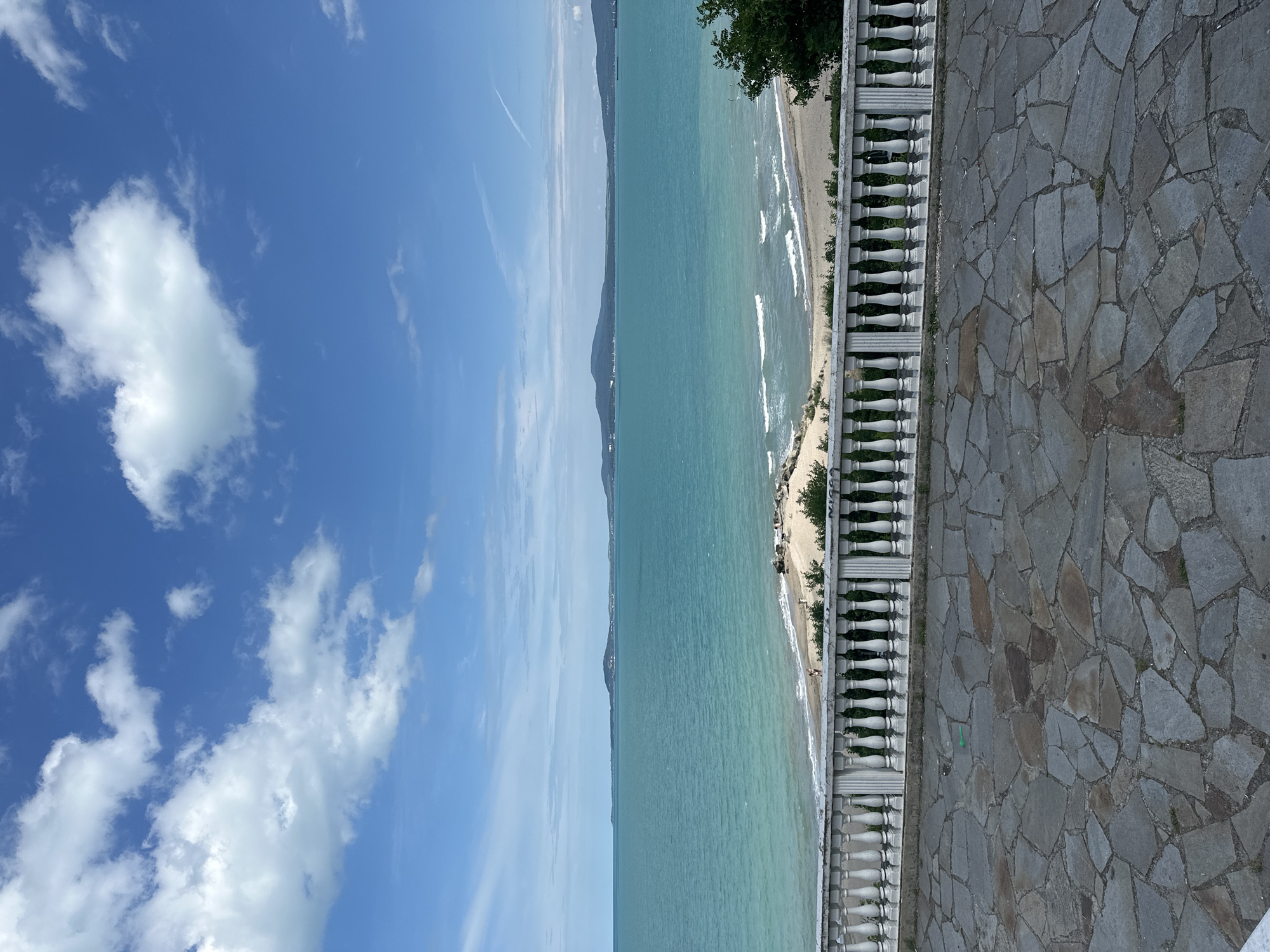 The image shows a view of the Black Sea from a paved terrace with a white balustrade in the foreground. The terrace is paved with irregular flagstones. Behind the balustrade, there is a sandy beach with a few scattered rocks and a line of green bushes and trees. The sea is a vibrant turquoise color with gentle waves lapping at the shore. In the distance, a hazy coastline with rolling hills can be seen. The sky is a clear blue with scattered white clouds, some of which are cumulus. The scene is likely a scenic overlook or promenade in Burgas, Bulgaria, offering a panoramic view of the coast. There are no people clearly visible in the image, and no discernable actions or interactions are taking place.