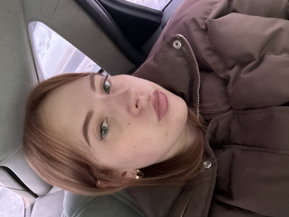 The image depicts a young woman seated within a vehicle, observed from the chest up. She has reddish-brown hair, green eyes, and wears pink-toned lipstick. A gold-colored hoop earring is visible on her left ear. She is dressed in a brown puffer jacket featuring a front zipper and snap buttons. Her gaze is directed towards the viewer. The vehicle's interior includes a grey fabric seat behind her and a visible headliner. Through the window to her right, a blurred, light-toned exterior environment, consistent with overcast or snowy conditions, is visible. The image contains no identifiable visual elements to confirm the specific location as Ol'khovochka, Russia.