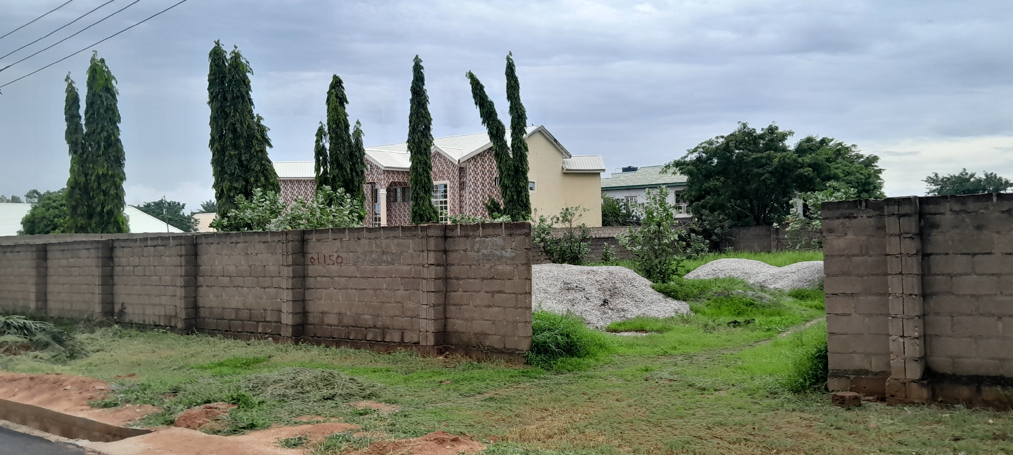 The image depicts a cloudy day scene in Jalingo, Nigeria, showcasing a boundary wall constructed from grey concrete blocks. The wall extends horizontally across the foreground, with a section of the wall in the mid-ground bearing the inscription "01150" in grey paint. Beyond the wall, on the right side, two large piles of light-colored gravel are visible on a patch of green grass. A narrow dirt path runs between the gravel piles and the wall. In the background, several tall, slender evergreen trees stand prominently. Behind the trees, a building with a white roof and brick-patterned walls is partially visible, with another building featuring a light yellow facade and a white roof situated to its left. Further in the distance, more trees and structures are present under a heavily overcast sky. The overall environment suggests an outdoor setting, possibly a construction site or a residential area undergoing development.