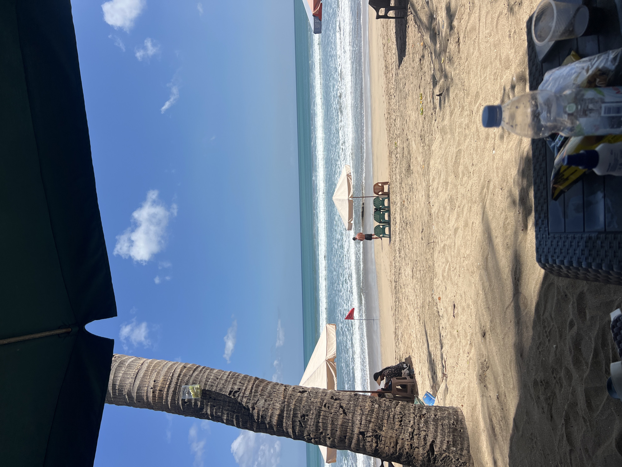 The image depicts a beach scene in Kuta, Indonesia, under a bright blue sky with scattered white clouds. In the foreground, the trunk of a large palm tree is prominent on the left, casting a shadow over the sandy beach. Partially visible umbrellas suggest a shaded seating area. Further back, a person sits at a small table under a beige umbrella. A red flag stands on the sand near the water's edge, and a man is visible standing on the beach. Several empty plastic chairs are arranged near another beige umbrella. The ocean features rolling waves, and the horizon is clearly defined. In the immediate foreground, a table is set with a plastic bottle of water, a clear plastic cup, and some wrapped items. The scene conveys a typical beach day atmosphere with elements of relaxation and leisure.