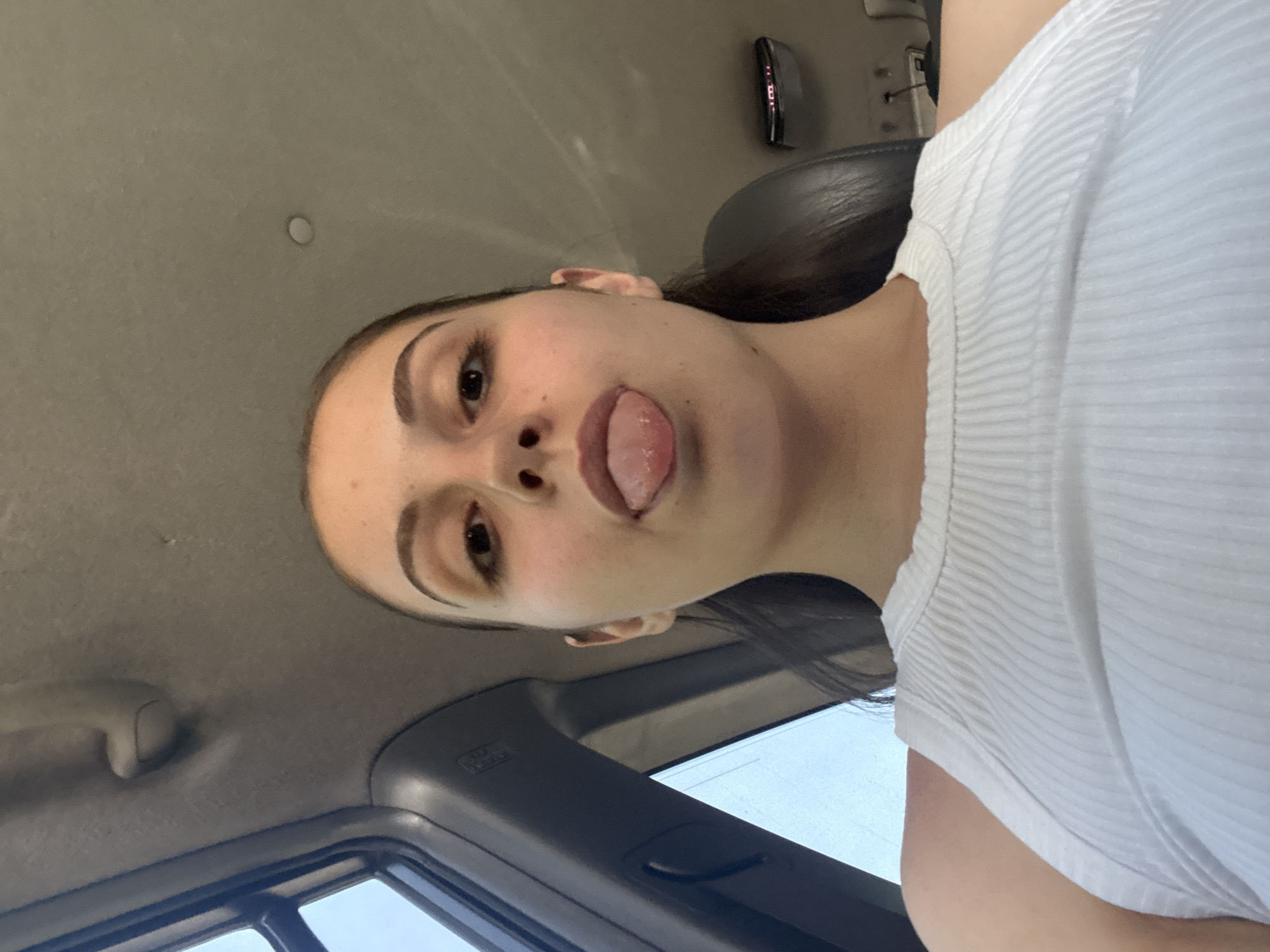A tilted, close-up photograph features a young woman inside a vehicle in Burgas, Bulgaria. She is seen from a low angle, with her head rotated to her left (viewer's right). She has dark hair pulled back, distinct eyebrows, and appears to be wearing light makeup. Her tongue is playfully sticking out slightly, covering her upper lip, as she looks directly at the camera.
She is wearing a white, ribbed, short-sleeved top. The background reveals the interior of a car or similar vehicle. Above her, the light gray headliner is visible, with a small, circular, light-colored object (possibly a light or sensor) and a dark console device or rearview mirror in the upper right. To her right, a dark, textured headrest and car seat are visible. Through the car window on the lower right, a bright, clear sky can be seen, indicating daytime. A dark frame around the window and a light gray passenger grab handle are also discernable. The text "AIRBAG" is visible on a panel near the window. The overall mood of the image is casual and playful.