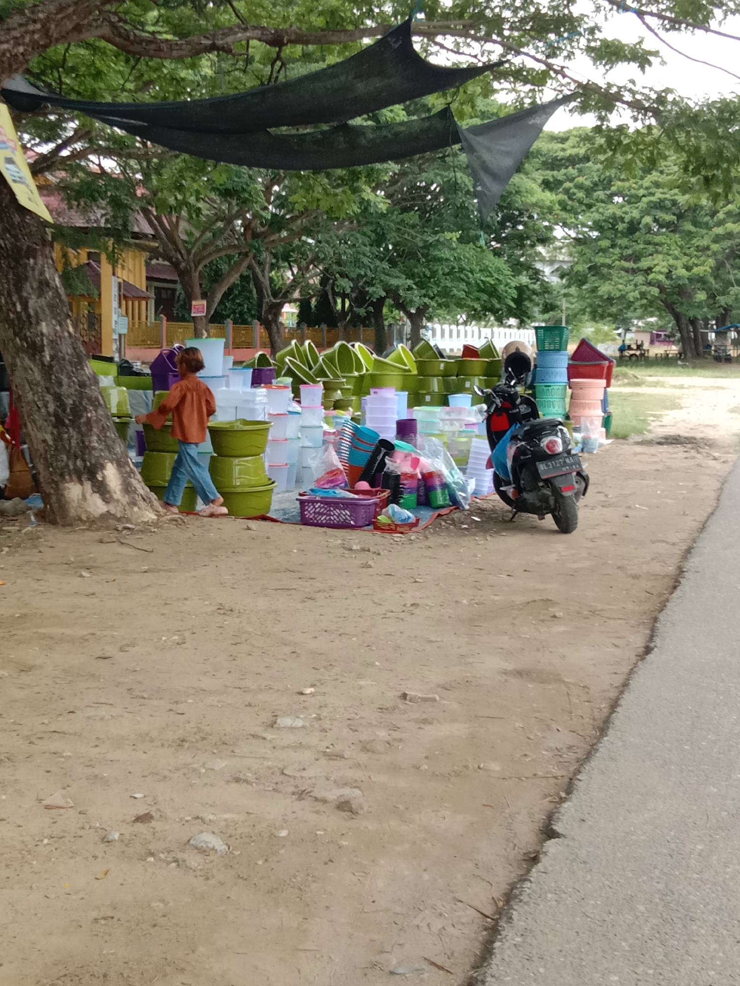The image depicts an outdoor market scene in Lhokseumawe, Indonesia, featuring a variety of plastic household items for sale. In the foreground, a dusty, unpaved area borders an asphalt road on the right. A large tree trunk with rough bark stands on the left, partially obscuring a yellow building in the background. A young person wearing an orange shirt and blue pants walks among stacks of green, clear, and purple plastic containers, bowls, and baskets. A black scooter with a license plate (BL 3127 NA) is parked among the merchandise. Overhead, dark netting is suspended from tree branches. The overall environment suggests a warm, possibly humid climate, with lush green trees visible in the background.